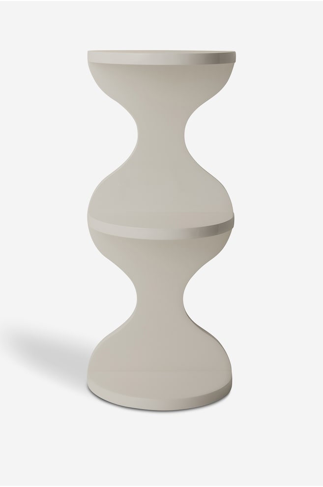 En ljusbeige skulptural piedestal stÃ¥r upprÃ¤tt och har en dubbel timglassilhuett med tre cirkulÃ¤ra nivÃ¥er. FÃ¶remÃ¥let har en slÃ¤t, matt finish med bÃ¶jda, symmetriska kolonner som fÃ¶rbinder den platta basen, den mellersta hyllan och den Ã¶vre ytan. - 1