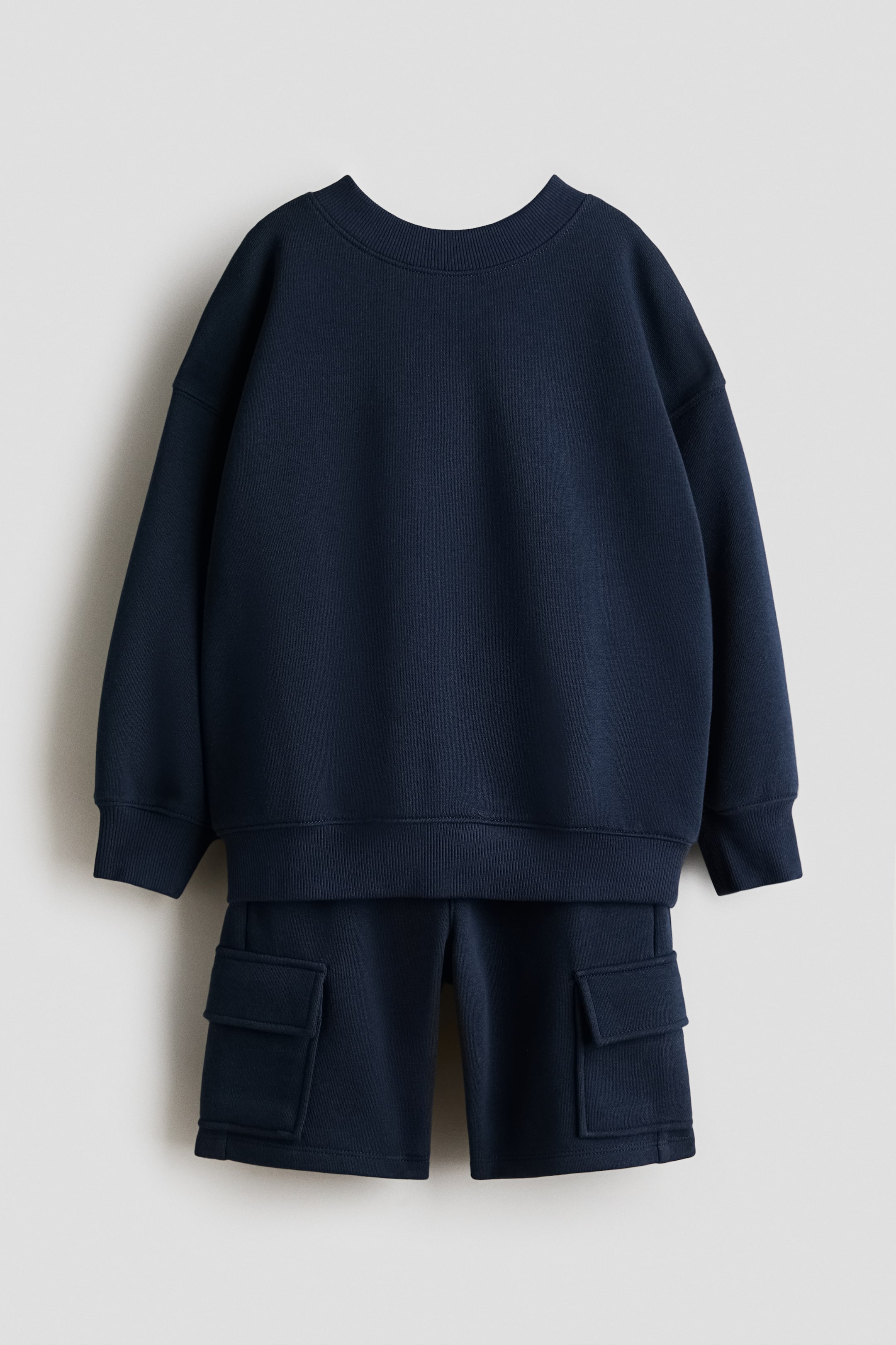 Ver imagem maior: A frente de um conjunto de sweatshirt azul‑marinho com calções combinantes está exibida. O sweatshirt apresenta uma gola redonda canelada, punhos canelados e uma barra canelada. Os calções têm dois grandes bolsos cargo na parte frontal de cada perna.