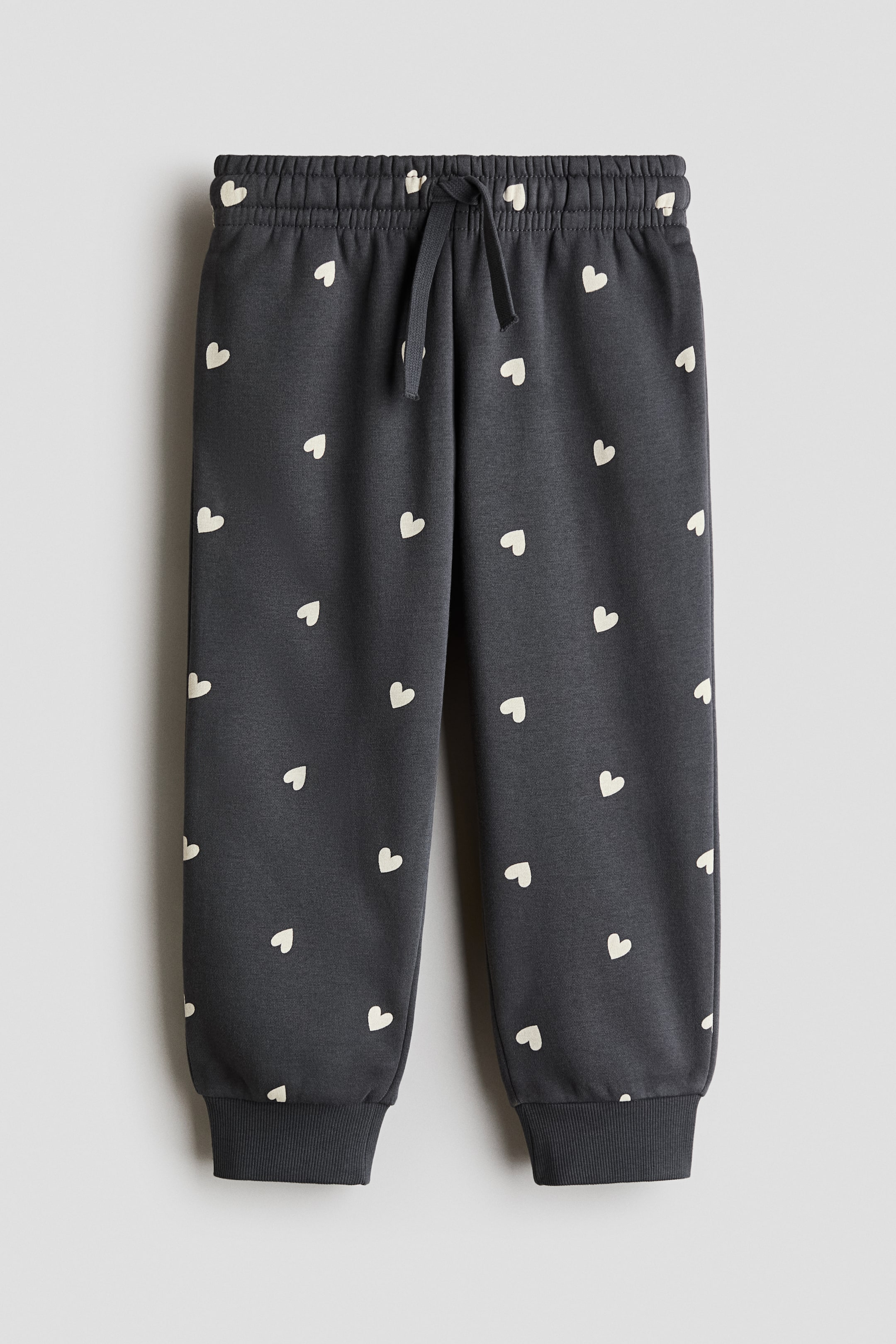 View larger image: Dark grey joggers are shown from the front, showcasing white heart motifs all over. They come with an elasticated drawstring waistband and ribbed cuffs at the ankles.