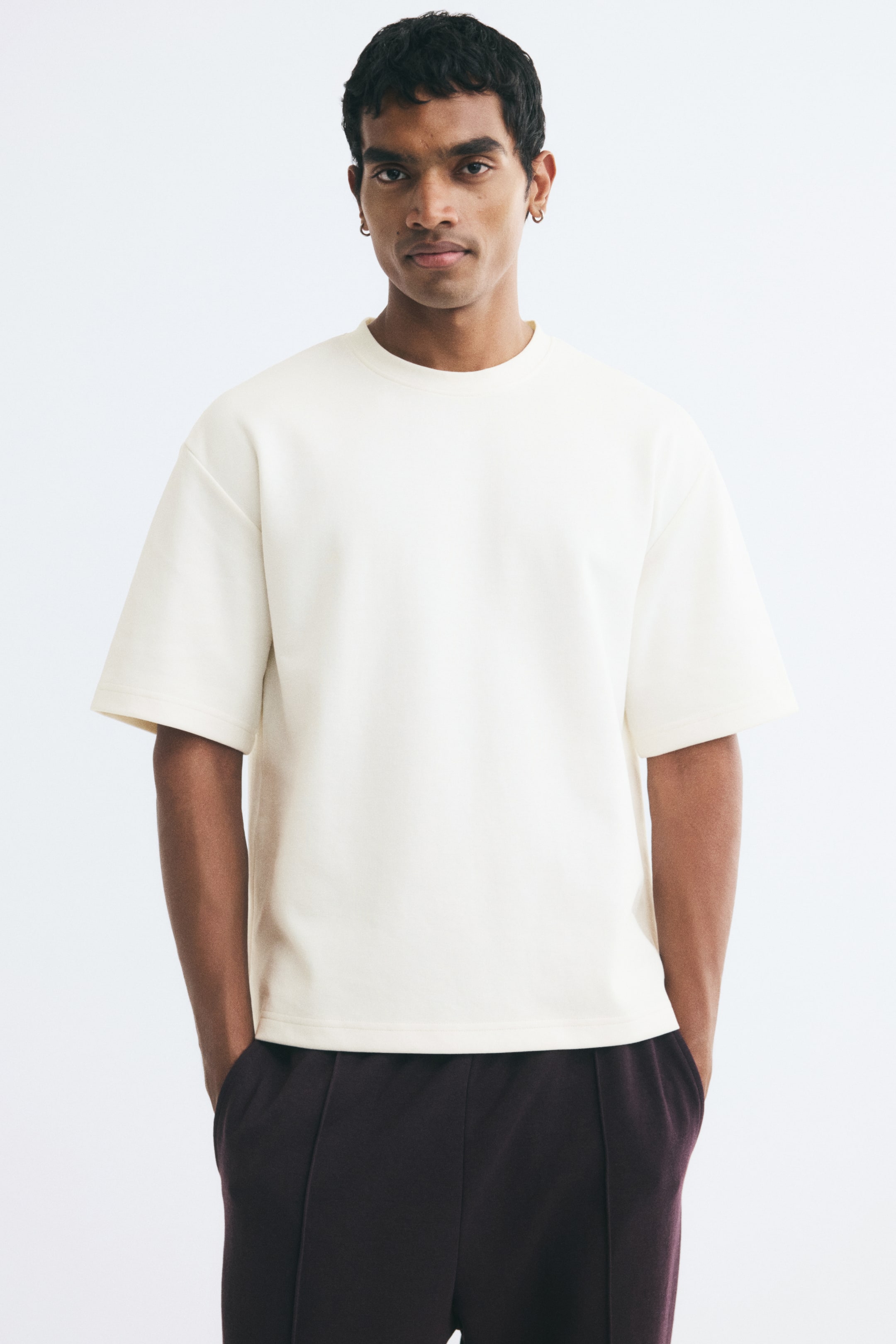 View larger image: A cream-coloured, heavyweight jersey tee with a relaxed fit, round rib-trimmed neckline, and dropped shoulders is being worn by the model. The tee features a straight hem and is paired with dark trousers, with the model standing with hands placed in their pockets.