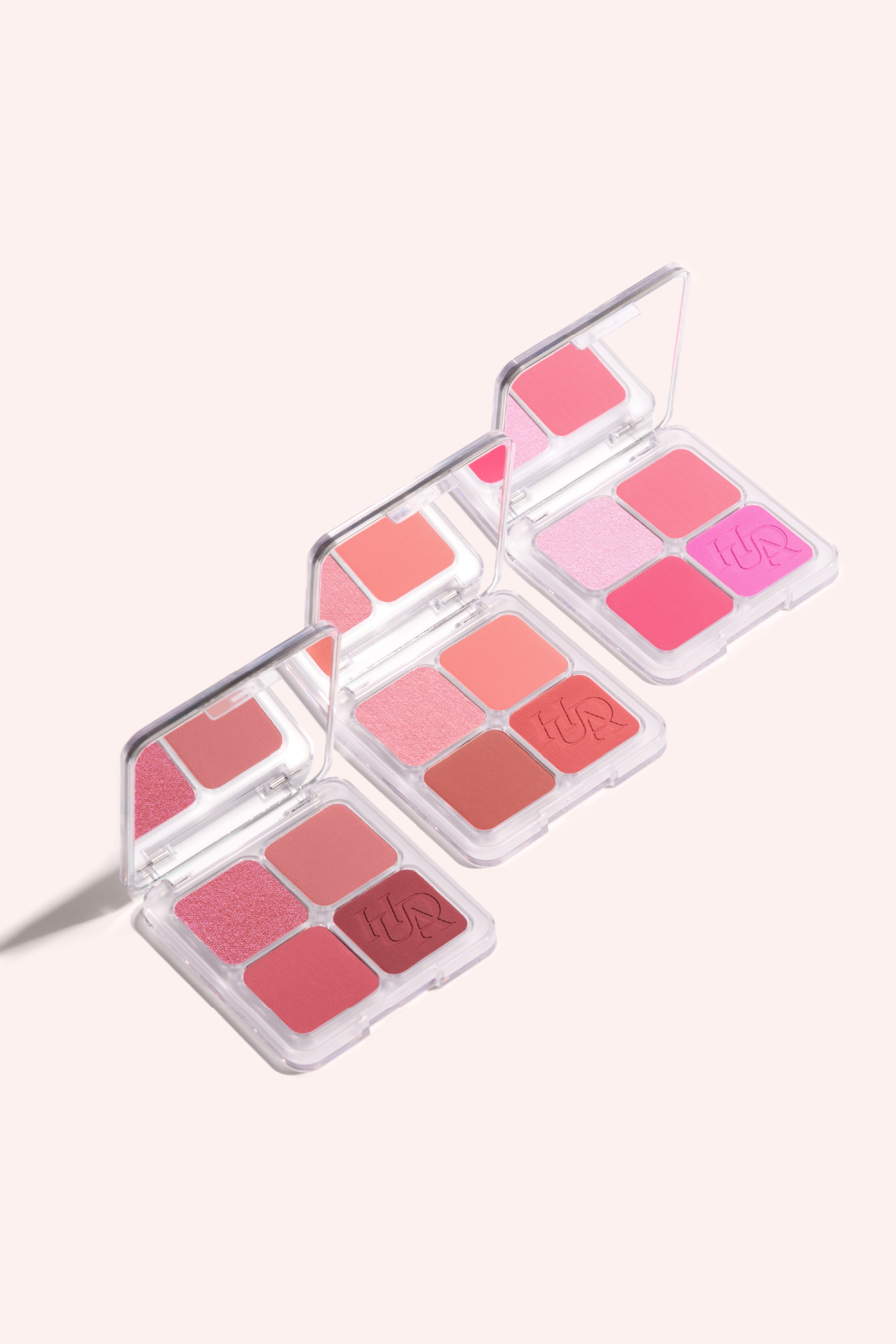 Vis større bilde: Tre klare plastikk paletter for sminke er arrangert diagonalt, hver med åpent lokk som avslører et speilet deksel og fire firkantede sminkepanner. Palettene viser frem forskjellige nyanser av rosa, korall og bærtoner, med noen panner som ser matte ut, andre skimrende, og én panne i hver palett har et preget logo.