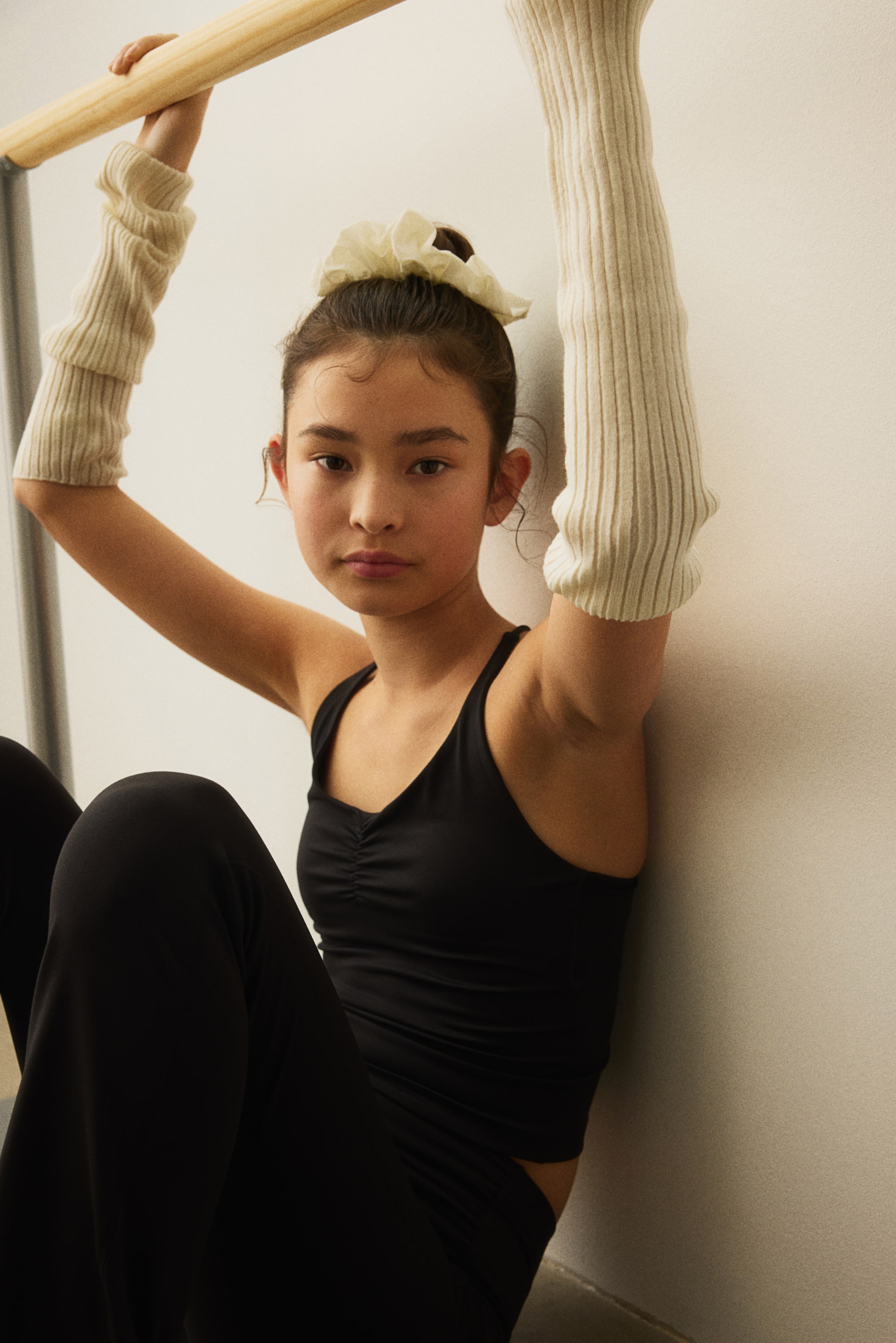 View larger image: A black camisole top features a V-neckline and central ruching, paired with black fitted trousers. Cream ribbed arm warmers cover the forearms, and a cream ruffled scrunchie is visible on the top of the head.