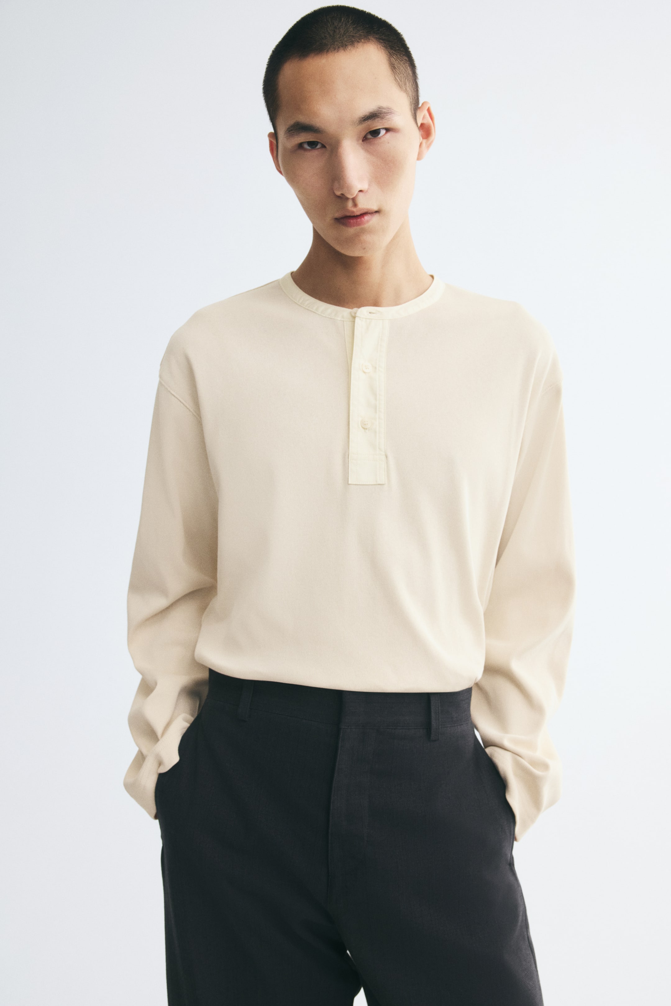 View larger image: The model is wearing a cream-coloured long-sleeve Henley shirt tucked into dark trousers. The shirt features a round neckline with a button placket, dropped shoulders, and a relaxed fit.