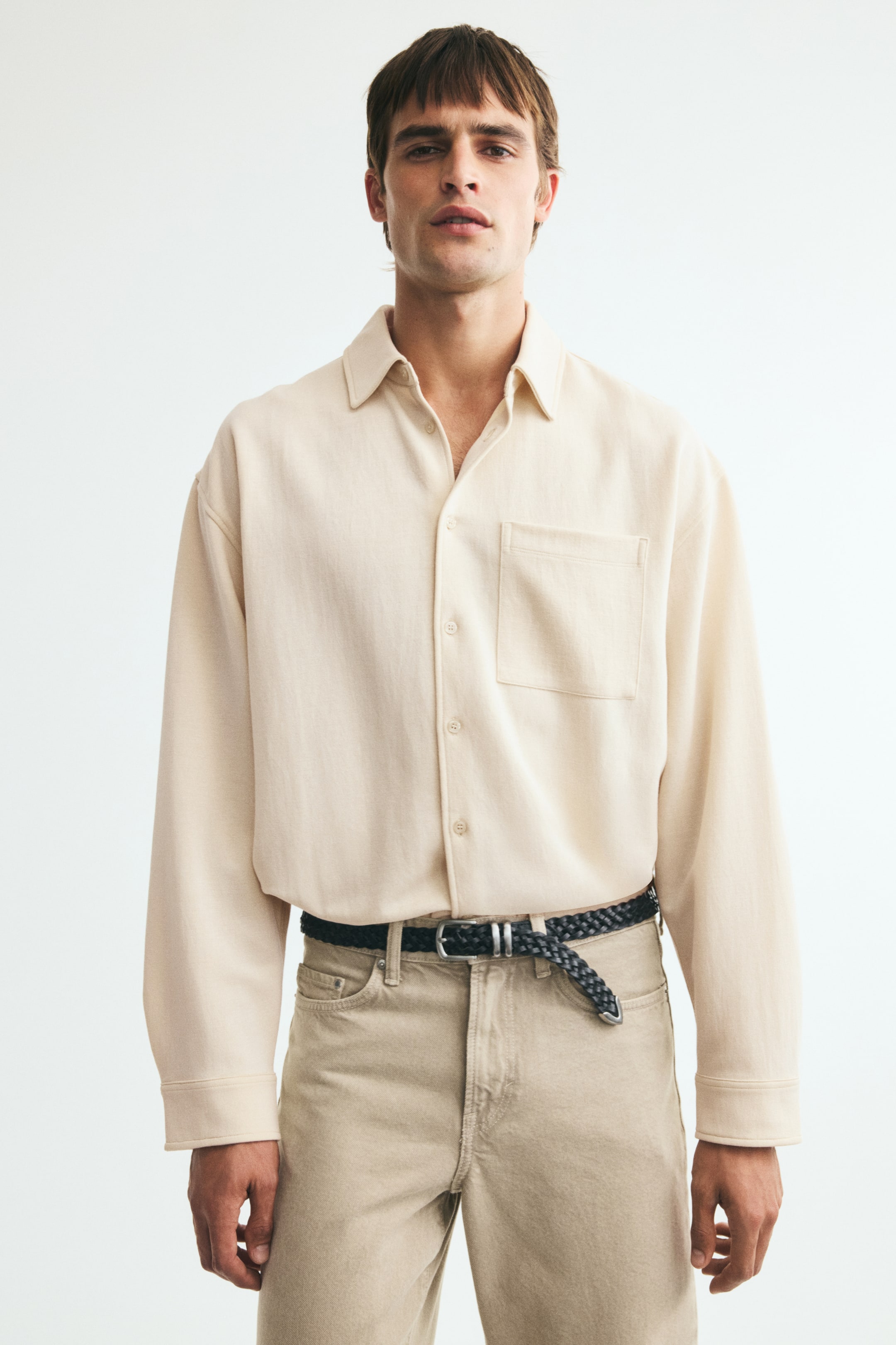 Xem ảnh lớn hơn: Một chiếc áo sơ mi interlock jersey màu be nhạt, có cổ áo, nẹp che nút kiểu French front, túi ngực mở, vai xuôi và tay dài với cổ tay cài nút, đang được người mẫu mặc. Chiếc áo sơ mi dáng relaxed-fit được sơ vin vào quần màu sáng.