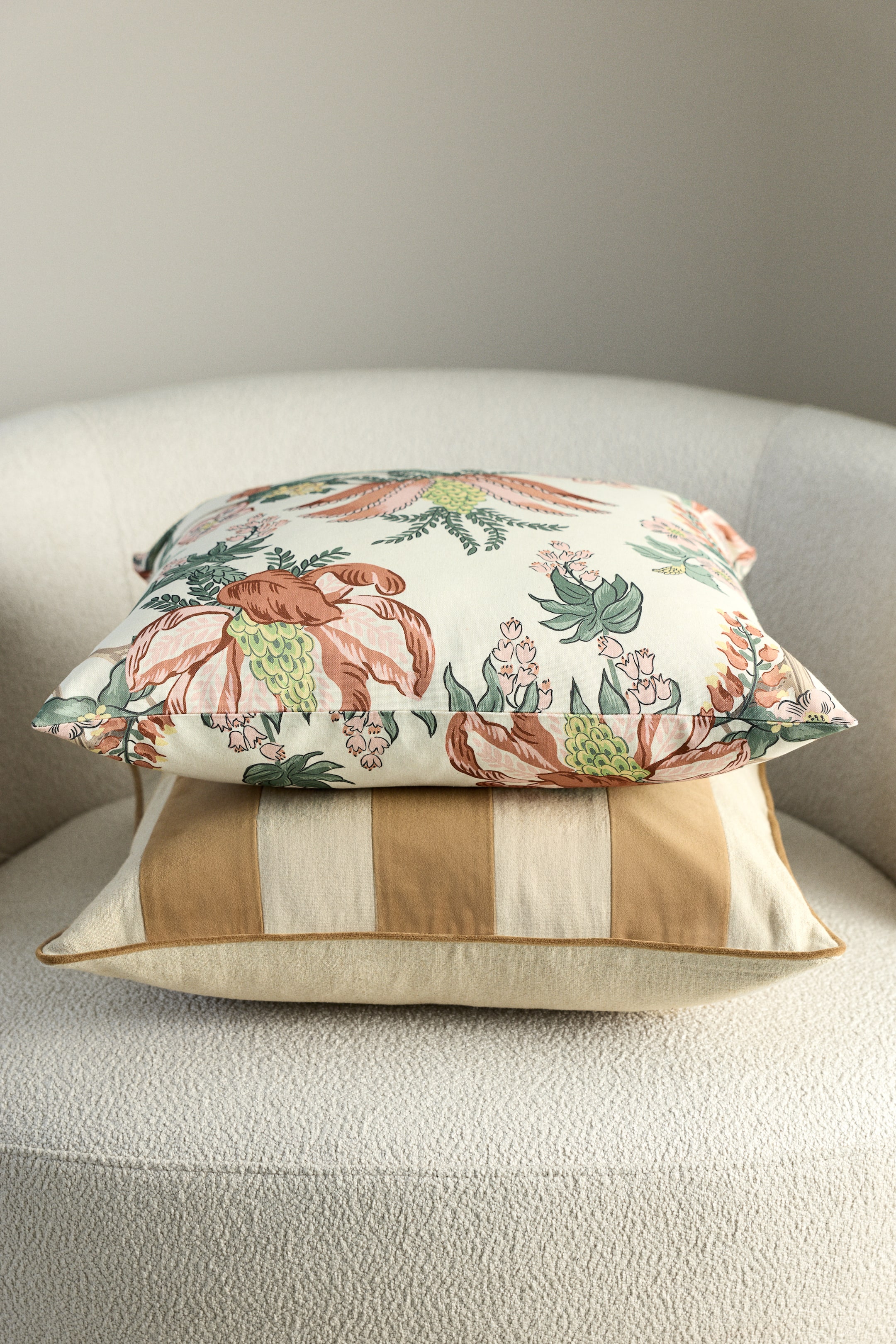 View larger image: Two cushions are stacked, one on top of the other. The top cushion is displaying a cream background with a multi-coloured floral pattern, showing large peach, green and yellow blooms. The bottom cushion is presenting alternating vertical stripes in cream and tan, and is finished with tan piping along its edges.