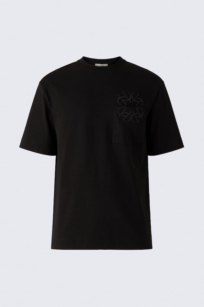 A front-facing view displays a black heavyweight cotton jersey t-shirt featuring a ribbed crew neckline, short sleeves, and a straight hem. A matching tonal embroidered monogram is overlapping the top edge of a single patch chest pocket. - 1