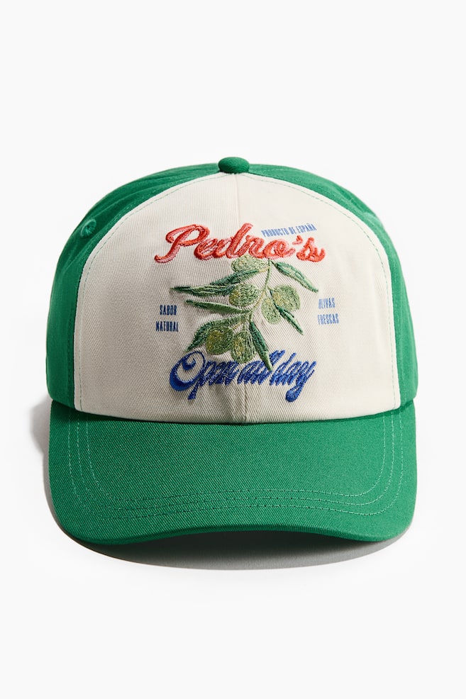 Une casquette en sergé de coton est orientée vers l'avant, avec un design bicolore comprenant des panneaux latéraux verts et une visière, ainsi qu'un panneau avant blanc cassé. Le panneau avant présente un motif brodé, incluant un texte rouge indiquant "Pedro's", une branche d'olivier avec des olives et des feuilles vertes, et un texte bleu indiquant "Ouvert toute la journée", accompagné de petites phrases en bleu comme "Producto de España", "Sabor Natural", et "Olivas Frescas." - 1