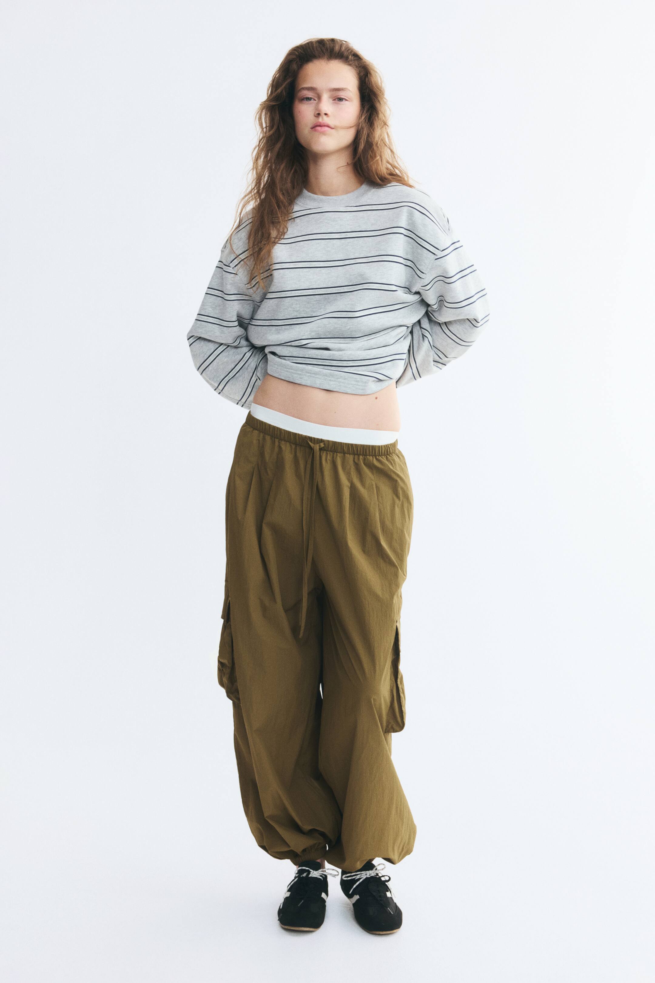 Ampliar la imagen: La modelo lleva puestos unos pantalones de nailon verde oliva de corte oversize, con cintura elástica con cordón ajustable, pinzas delanteras, bolsillos laterales tipo fuelle y bajos fruncidos con elástico. Los pantalones se combinan con una sudadera gris a rayas y tenis negros.