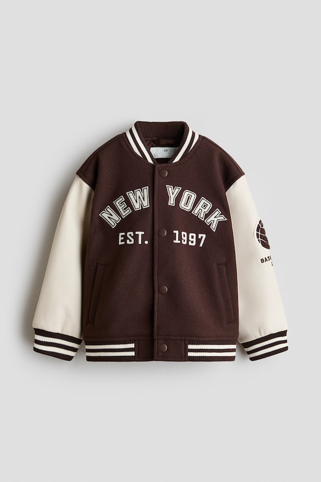 Eine Baseballjacke wird von vorne präsentiert, mit einem dunkelbraunen, gefilzten Körper und kontrastierenden, cremefarben beschichteten Ärmeln. Auf der Vorderseite der Jacke steht "NEW YORK EST. 1997" in cremefarbener Schrift, und auf dem rechten Ärmel ist ein Basketballmotiv zu sehen. Sie ist mit einem gerippten Kragen, breiten gerippten Bündchen und einem breiten gerippten Saum in Dunkelbraun mit cremefarbenen Streifen gestaltet und verfügt über dunkelbraune Druckknöpfe und Leistentaschen an der Vorderseite. - 1