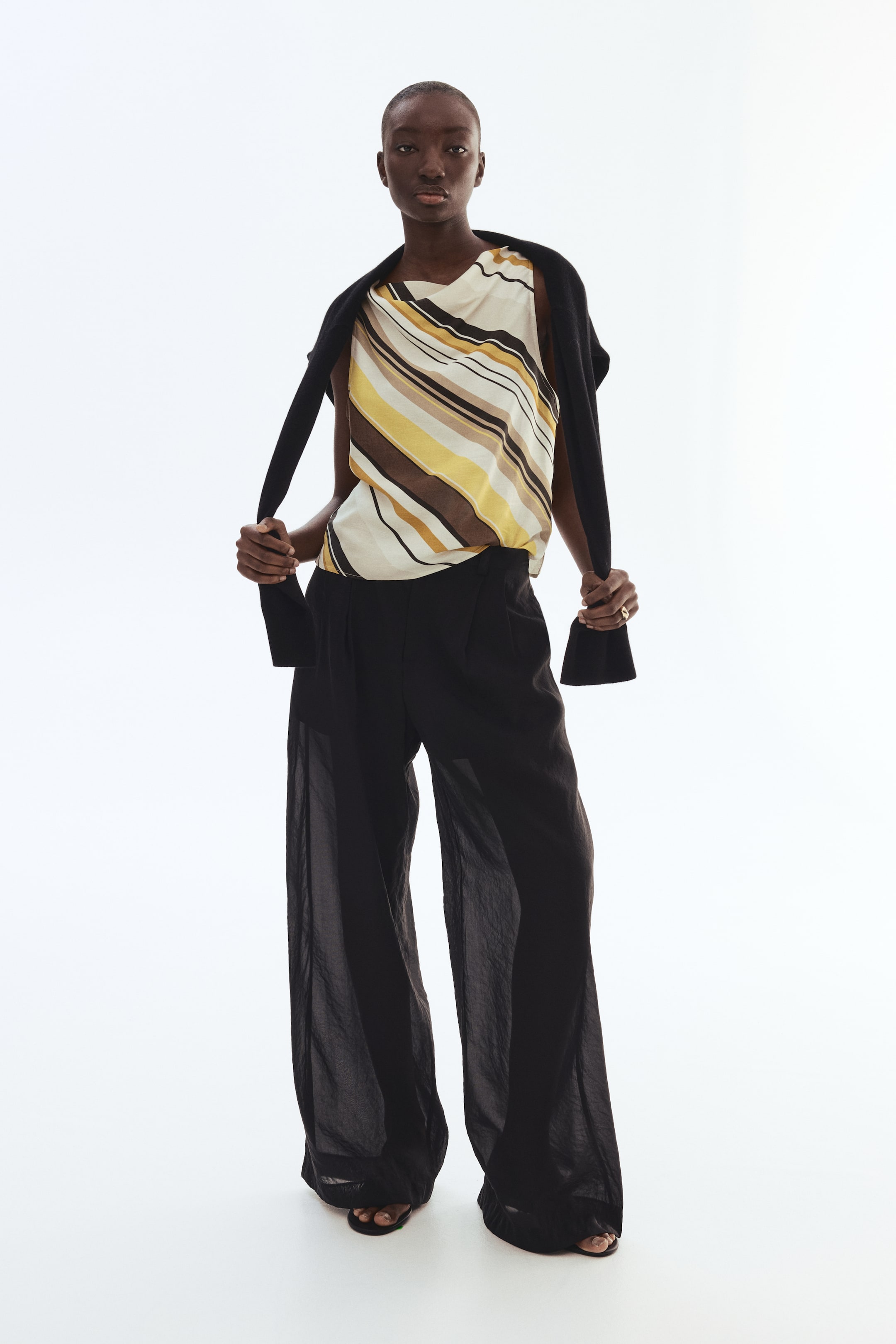 View larger image: The model is wearing a sleeveless top with a cowl neckline, showcasing a diagonal striped pattern in shades of cream, yellow, brown, and black. She is holding the ends of a black sweater that is elegantly draped over her shoulders.