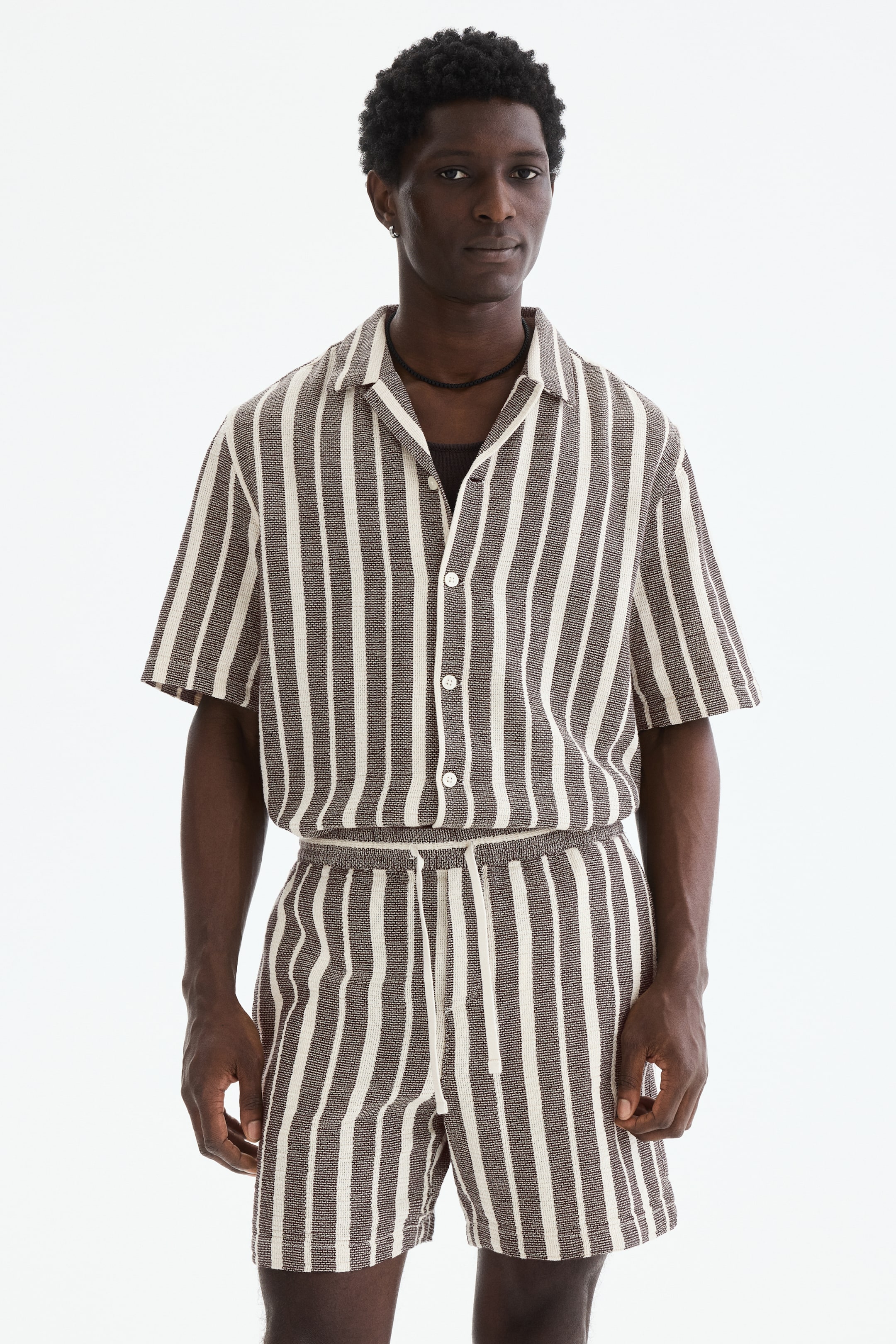 Voir une image plus grande: Un ensemble assorti de chemise à manches courtes et de shorts est présenté de face, avec des rayures verticales texturées marron et blanc cassé. La chemise est conçue avec un col à revers et une fermeture à boutons blancs, révélant un sous-vêtement foncé, tandis que le short présente une taille élastiquée avec cordon de serrage.