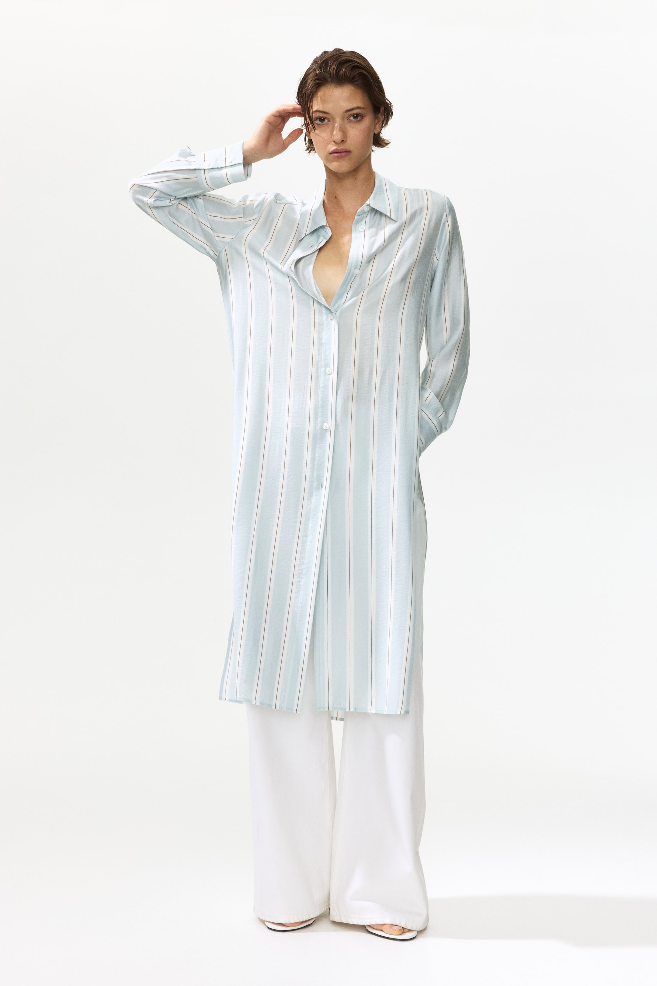 View larger image: "A light blue longline button-down shirt features thin white and tan vertical stripes. It comes with a pointed collar, long sleeves, and a partially unbuttoned front forming a V-neck opening, while its flowing silhouette extends over white wide-leg trousers."