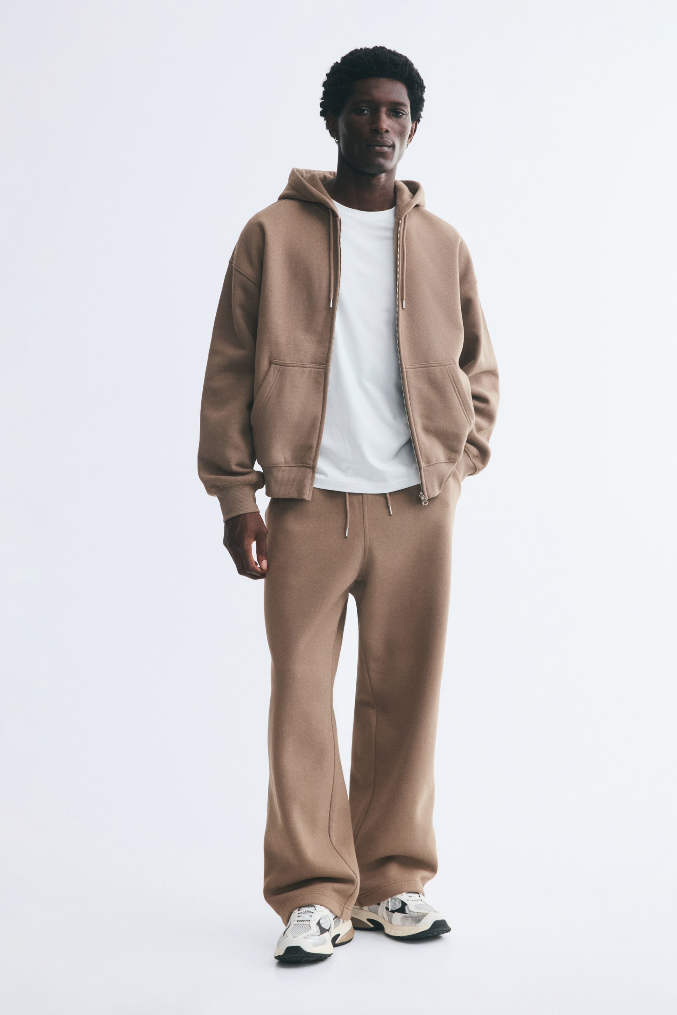 Agrandir l'image: Le mannequin porte un pantalon de survêtement en tissu molletonné mélangé de coton marron clair, avec une coupe ample et une taille élastiquée avec cordon de serrage. Le pantalon est associé à un hoodie zippé ouvert assorti, un t-shirt blanc et des baskets claires.