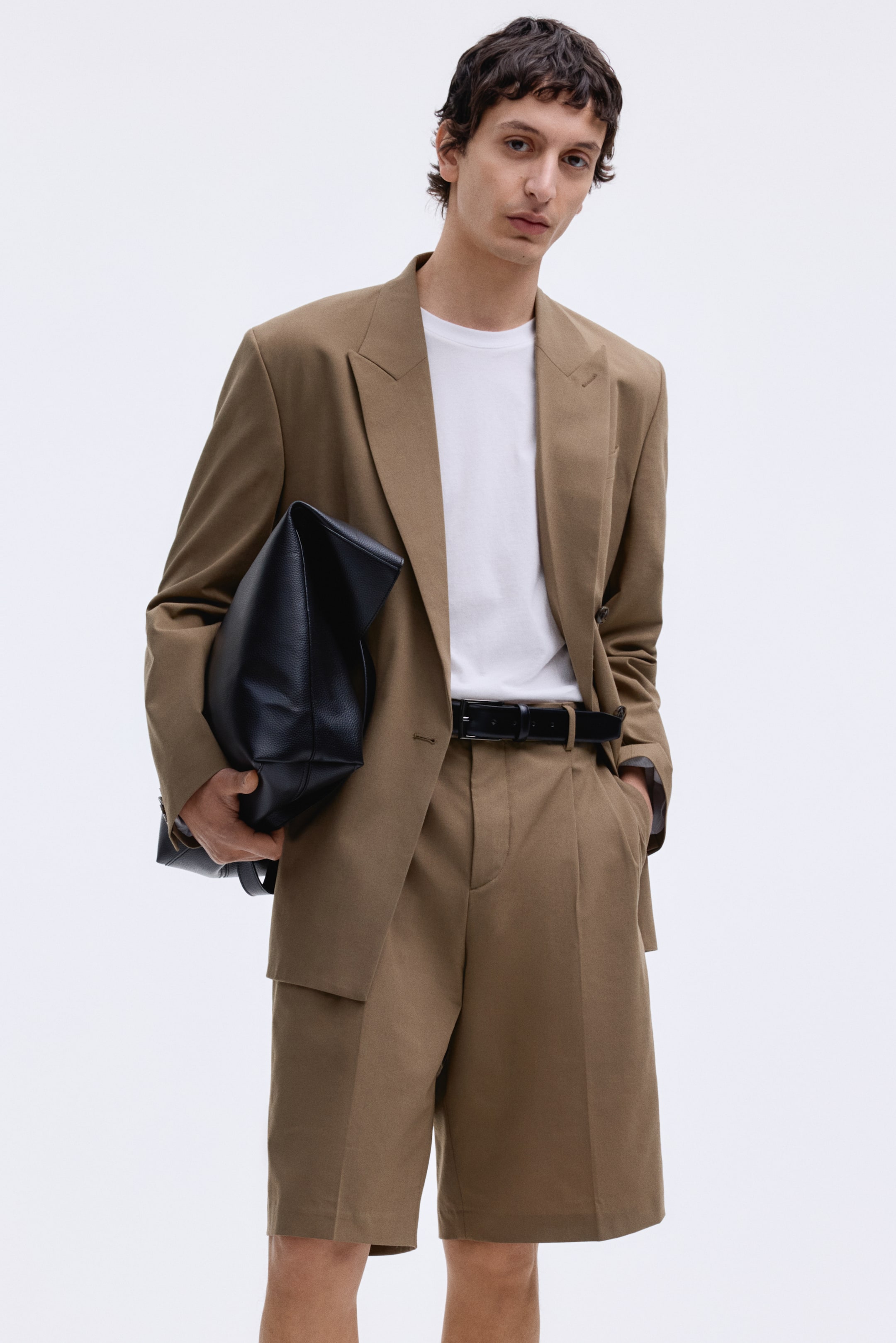 Grotere afbeelding bekijken: Een op maat gemaakt zandkleurig pak, bestaande uit een enkelrij blazer met inkeping revers en klepzakken en bijpassende geplooide shorts die boven de knie eindigen, wordt gepresenteerd met de voorkant naar voren. De blazer wordt gedragen over een wit t-shirt, en de shorts zijn vastgemaakt met een zwarte riem, terwijl een zwarte gestructureerde tas tegen de blazer wordt gehouden.