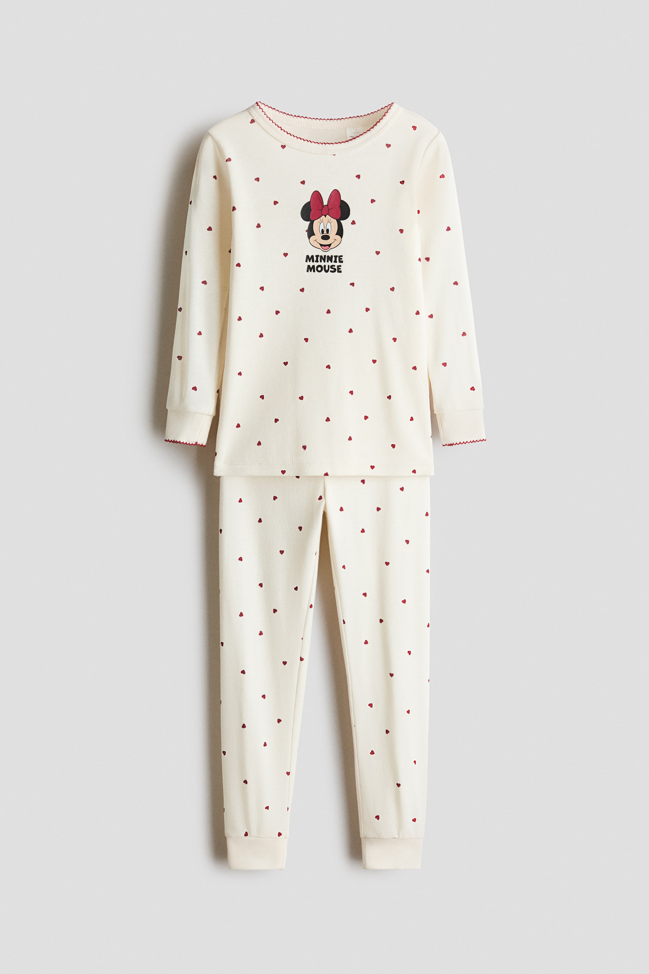 View larger image: This front view shows a cream-colored cotton jersey pajama set featuring an all-over tiny red heart pattern on both the long-sleeve top and matching pants. The top displays a Minnie Mouse graphic on the chest with red picot trim at the neckline and cuffs, while the pants feature an elasticized waist and cuffed ankles.