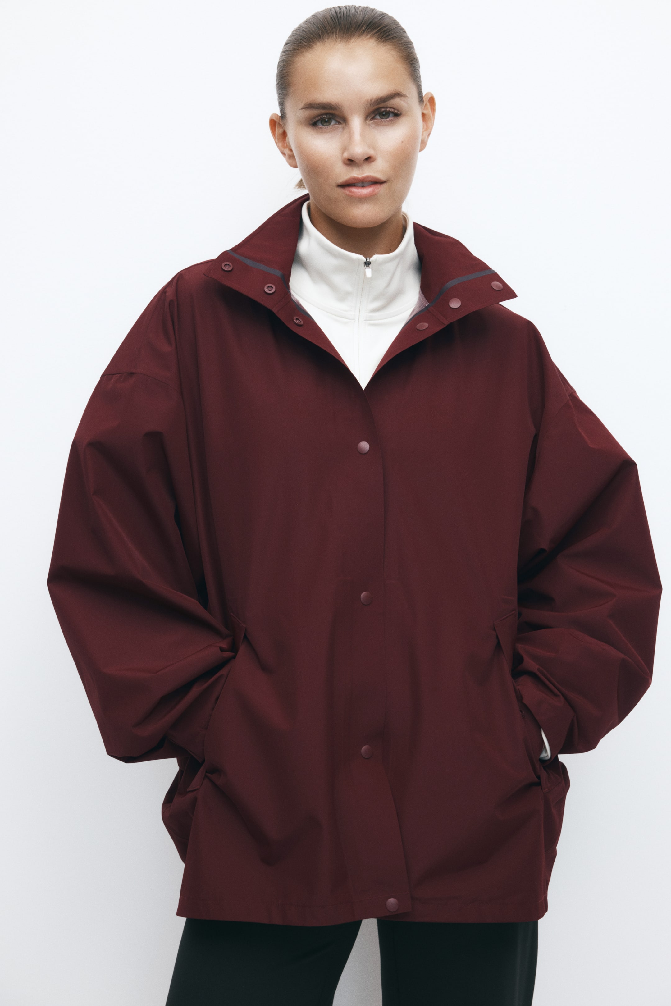 Se større billede: En bordeauxfarvet oversized jakke med trykknapper foran og en bred krave bæres af modellen, som står med hænderne i sidelommerne. Jakken er stylet over en hvid, højhalset top med lynlås.
