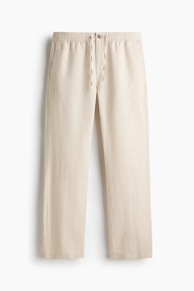 Light beige linen pants are displaying an elasticated waistband with a visible drawstring and a single button closure. They are featuring slanted side pockets and a relaxed straight-leg cut. - 1