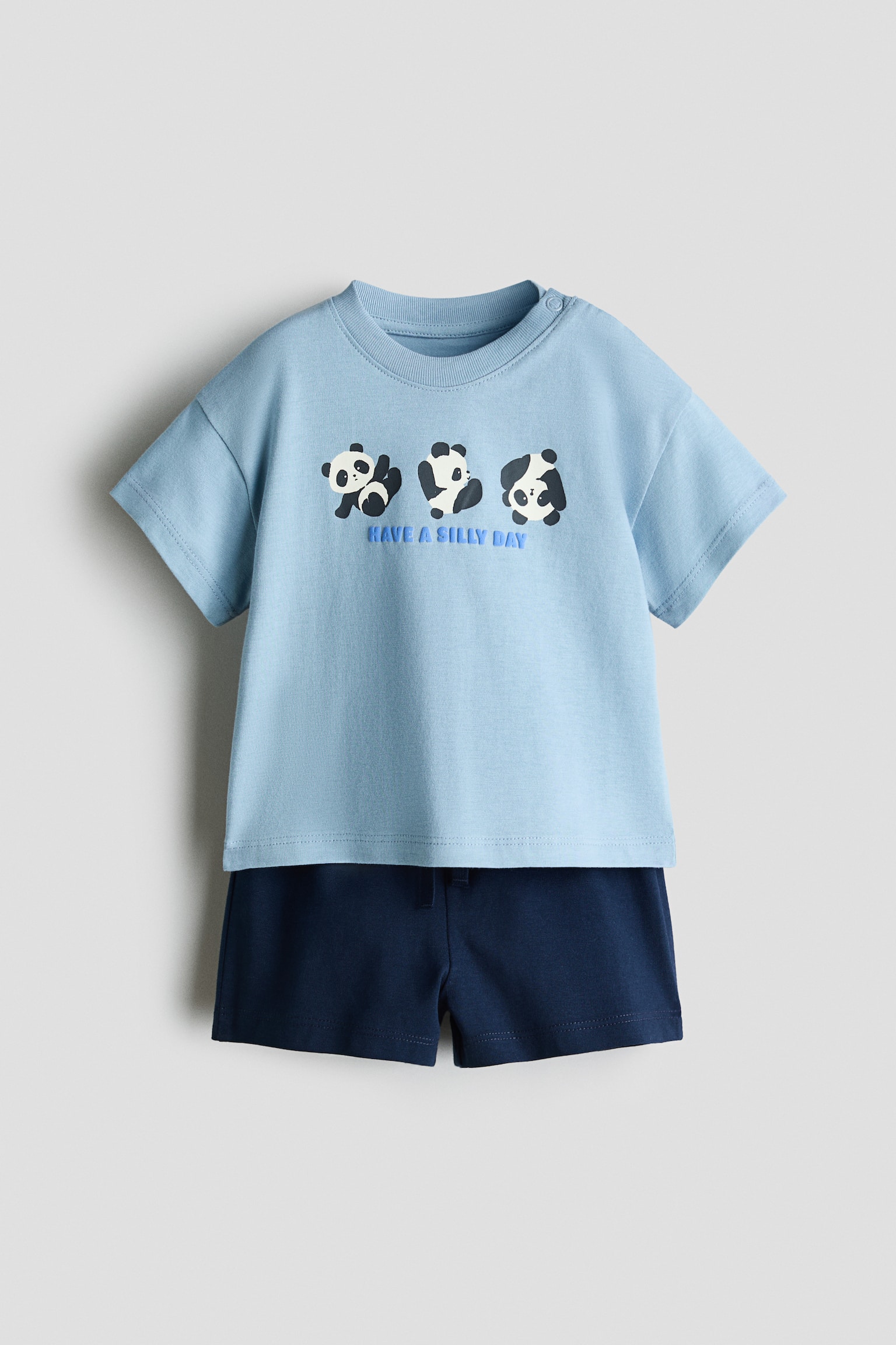 Má»t bá» cotton 2 món ÄÆ°á»£c chụp từ phÃa trưá»c, gá»m áo thun tay ngắn mà u xanh da trá»i nhạt và quần short mà u xanh navy Äáºm. Ão thun in há»a tiết ba chú gấu trúc hoạt hình phÃa trên dòng chữ "HAVE A SILLY DAY" á» ngá»±c, trong khi quần short Äá»ng bá» có cạp chun co giãn.