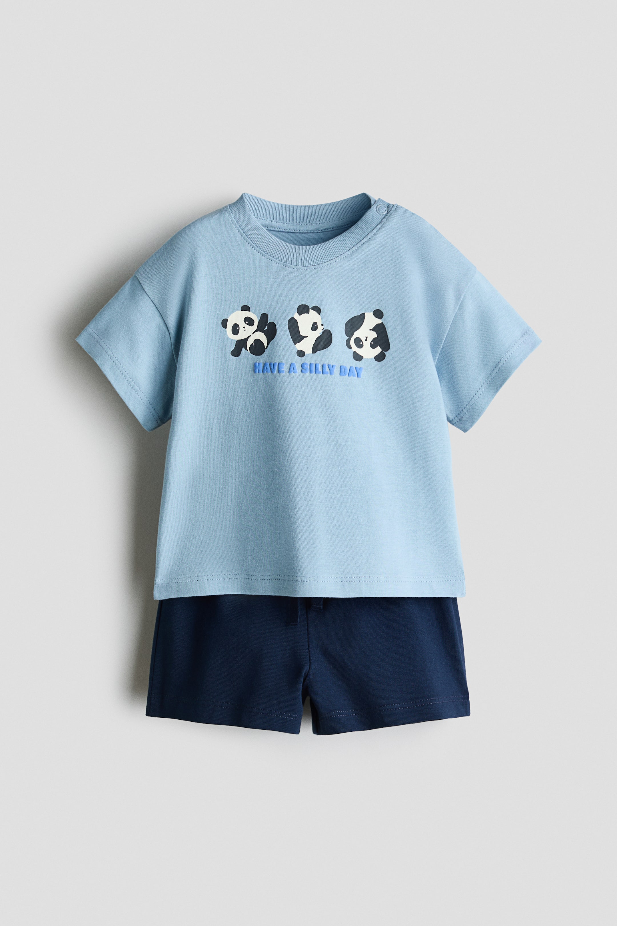 View larger image: A 2-piece cotton set is shown from the front, featuring a light blue, short-sleeved T-shirt and dark navy blue shorts. The T-shirt displays a graphic of three cartoon pandas above the text "HAVE A SILLY DAY" on the chest, while the matching shorts feature an elasticized waistband.