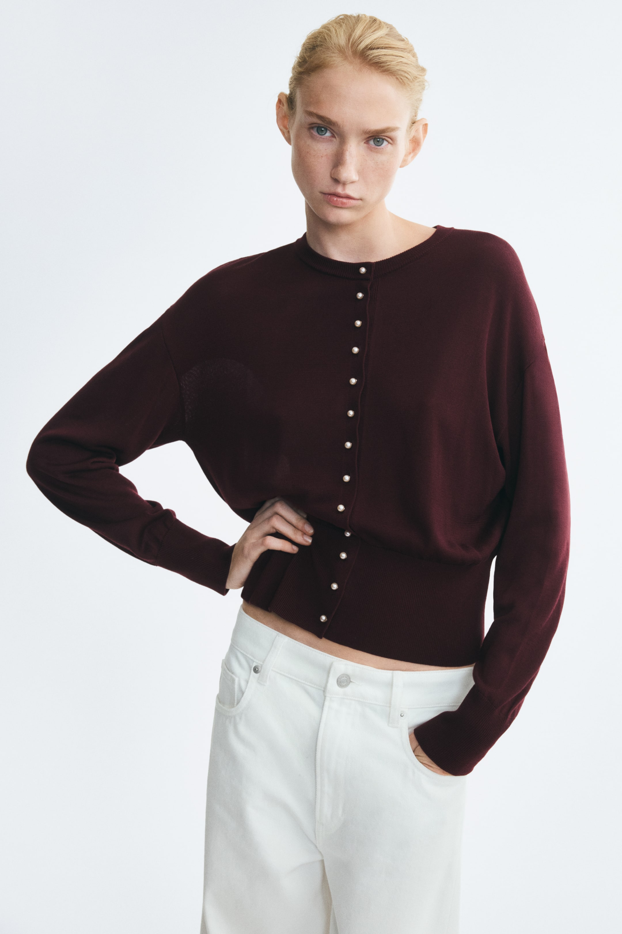 View larger image: A burgundy knit cardigan featuring a round neckline, pearly buttons down the front, dropped shoulders, and a wide ribbed waistband is being worn by the model. The model is pairing the cardigan with white pants, posing with one hand on their hip and the other in a pocket.