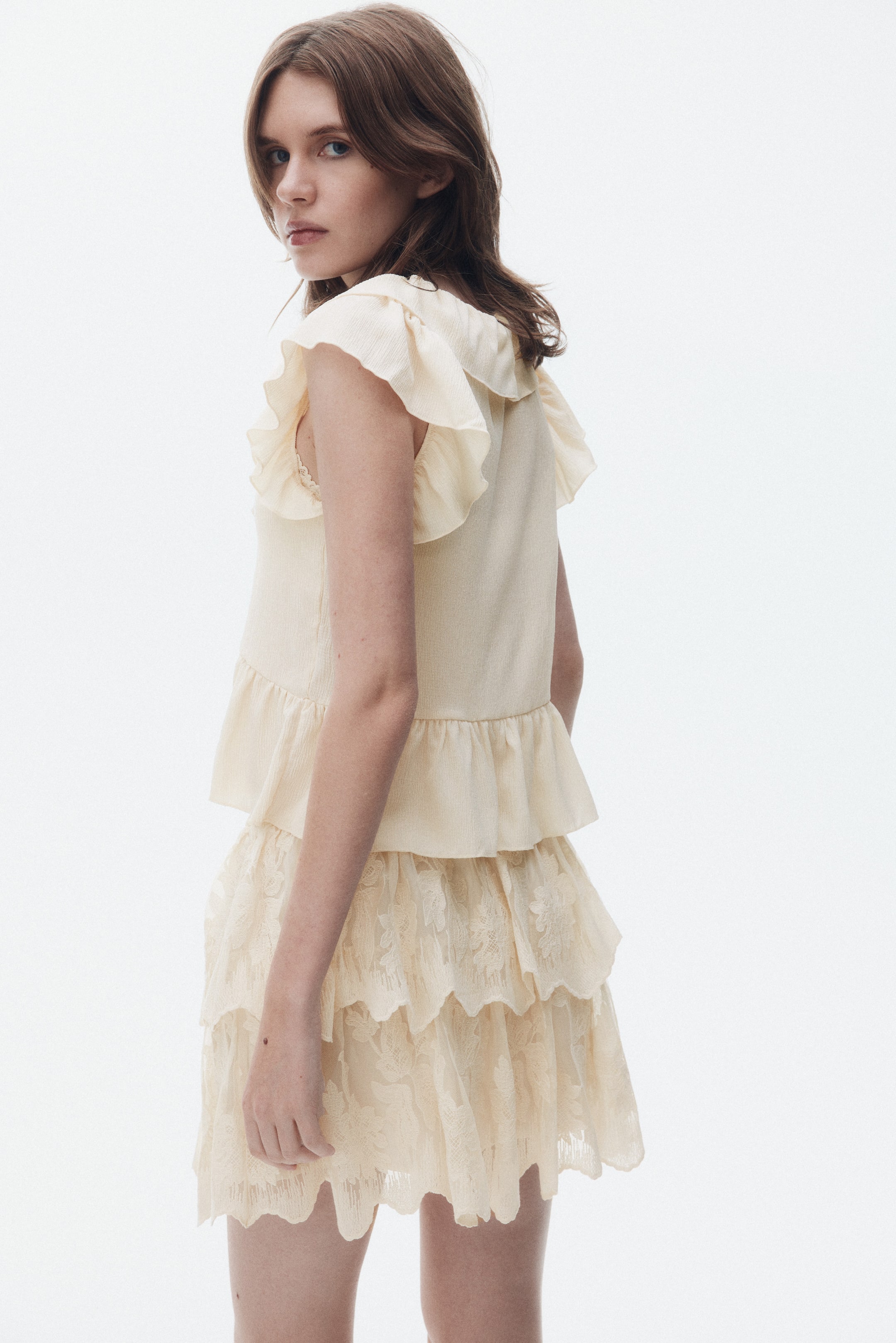 Ampliar la imagen: Un conjunto de blusa y falda color crema se presenta desde un ángulo trasero izquierdo, mostrando los volantes de la blusa a lo largo de los hombros y el dobladillo peplum con volantes. La falda está diseñada con múltiples capas escalonadas de tela similar al encaje, exhibiendo patrones florales bordados y un borde festoneado.