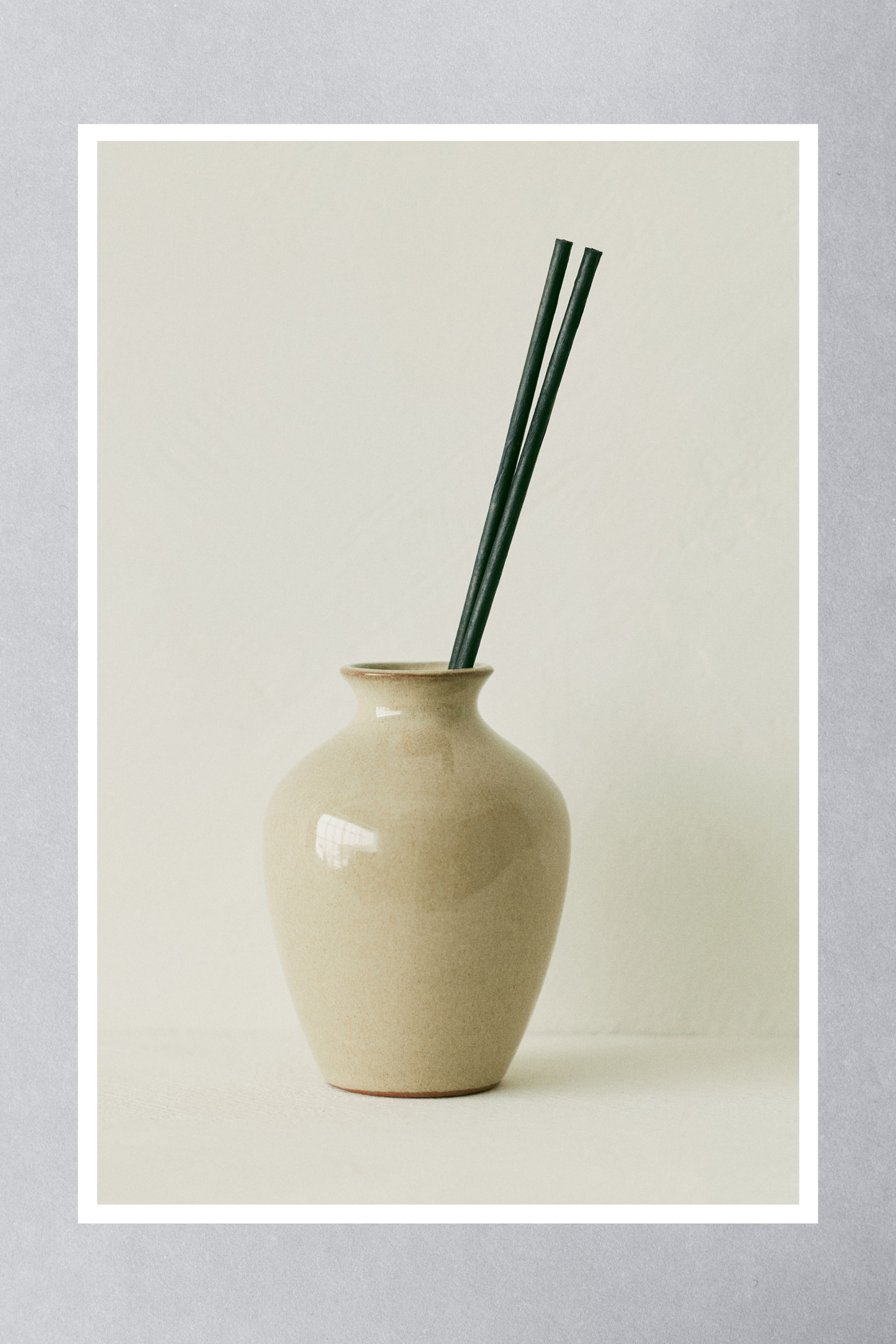 Ingrandisci l'immagine: Due bastoncini profumati di carta verde scuro si ergono da un vaso in ceramica beige chiaro, inclinati leggermente verso destra.