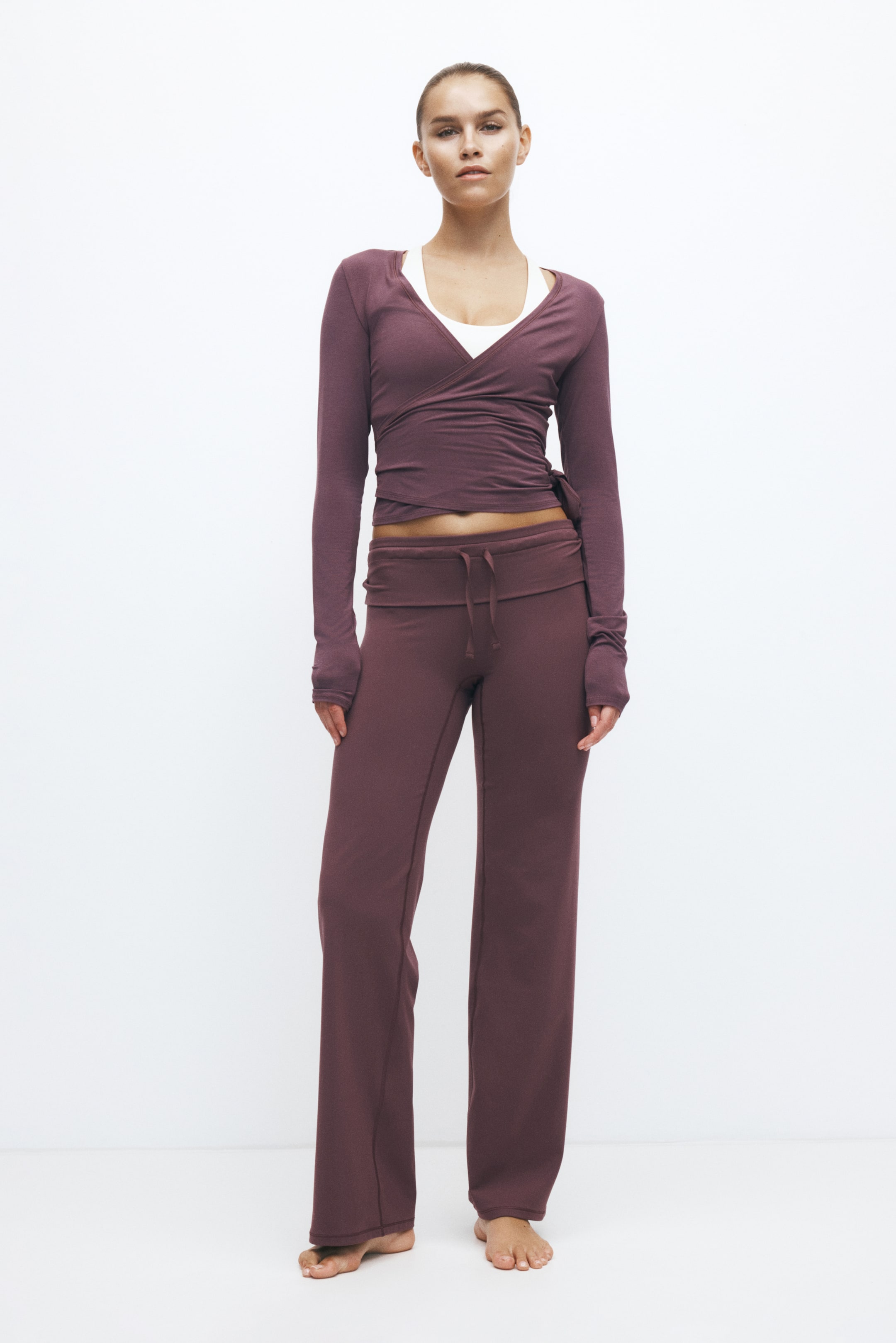 Zobrazit větší obrázek: Modelka má na sobě loungewear soupravu v odstínu mauve z žebrovaného úpletu, která se skládá z dlouhého zavinovacího topu s rukávy a ladících širokých kalhot. Zavinovací top je v pase uvázaný na boku přes bílé tílko, zatímco kalhoty mají přehnutý pas se stahovací šňůrkou.
