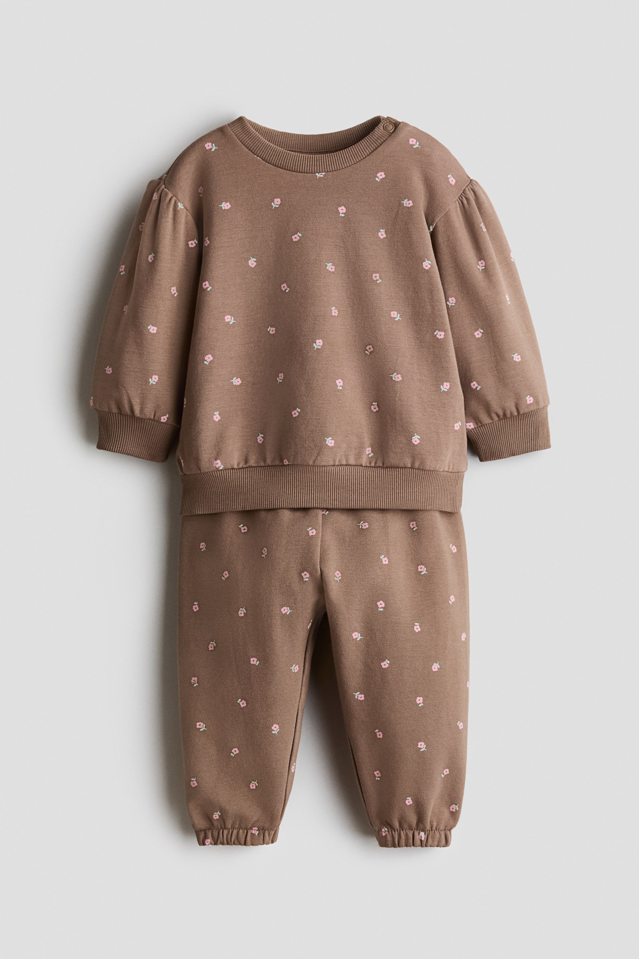 View larger image: A brown two-piece set features a small pink floral print throughout. The long-sleeved top features a round neck, a snap button closure on one shoulder, puffed sleeves, and ribbed cuffs and hem, while the matching pants are designed with elasticated cuffs.