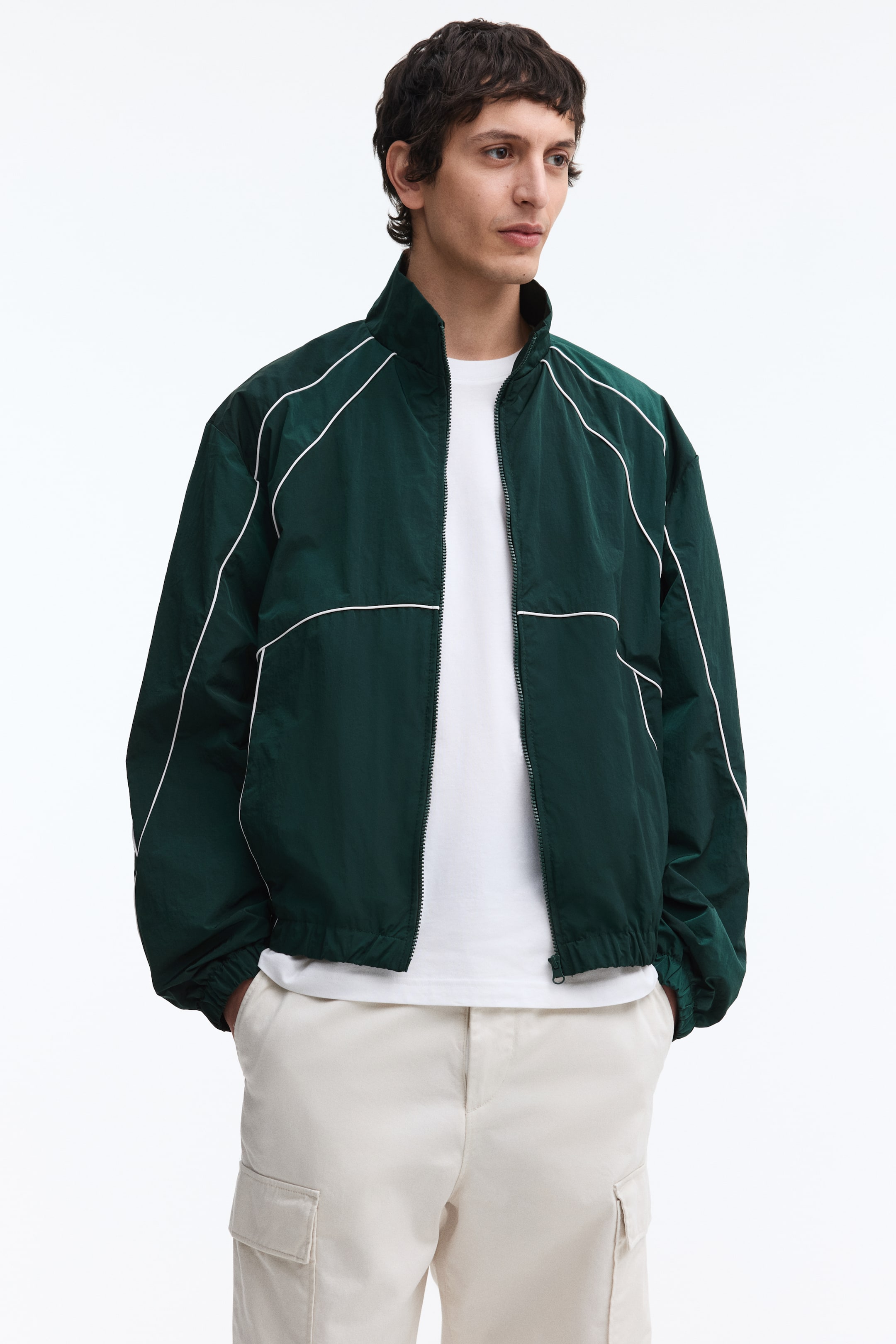 View larger image: A dark green track jacket featuring white piping details along the shoulders, chest, and arms, with a stand-up collar, a full-length front zipper open over a white t-shirt, and elasticated cuffs and hem.