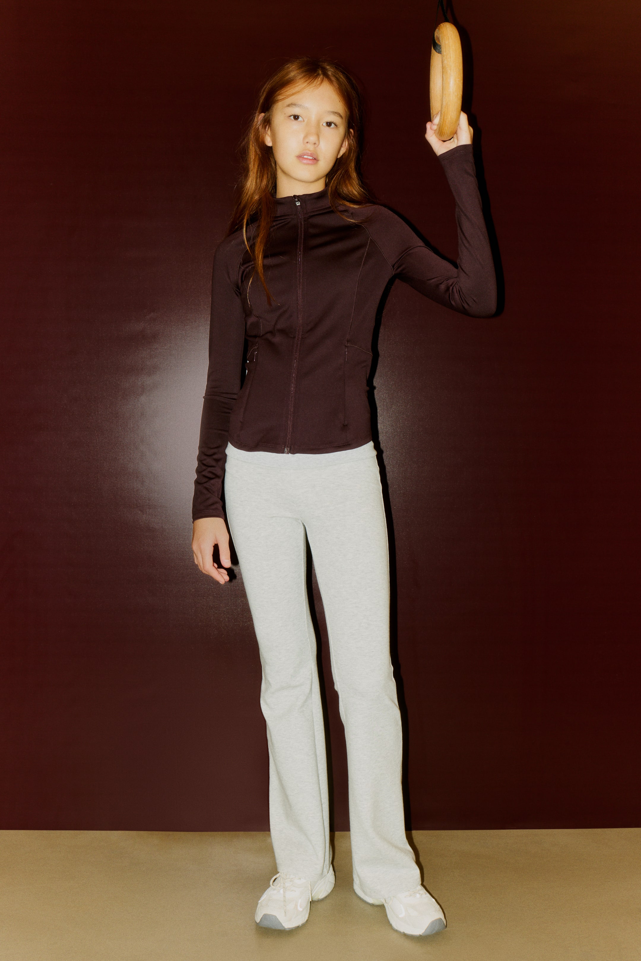 Ampliar la imagen: Una chaqueta deportiva ajustada de color marrón oscuro, con un cierre completo en la parte delantera, un cuello alto, mangas largas y paneles laterales visibles, se presenta de FRENTE. Se lleva con pantalones de chándal de corte de bota en gris claro, que cuentan con una cintura ajustada y una textura suave.