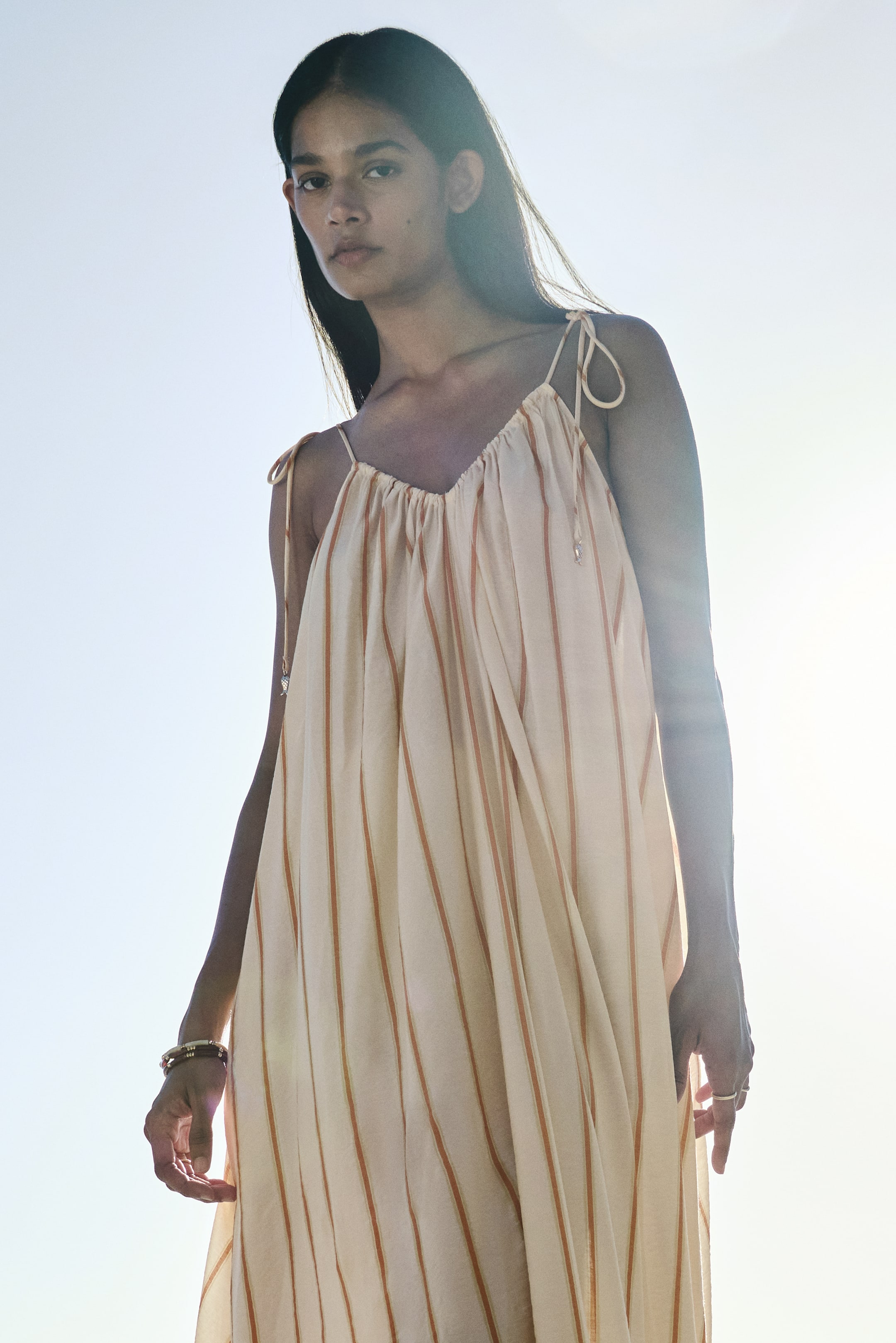 View larger image: An off-white dress with thin vertical orange stripes features a gathered V-neckline and tied spaghetti straps at the shoulders, showcasing its loose, flowing fit and drape from a low angle.