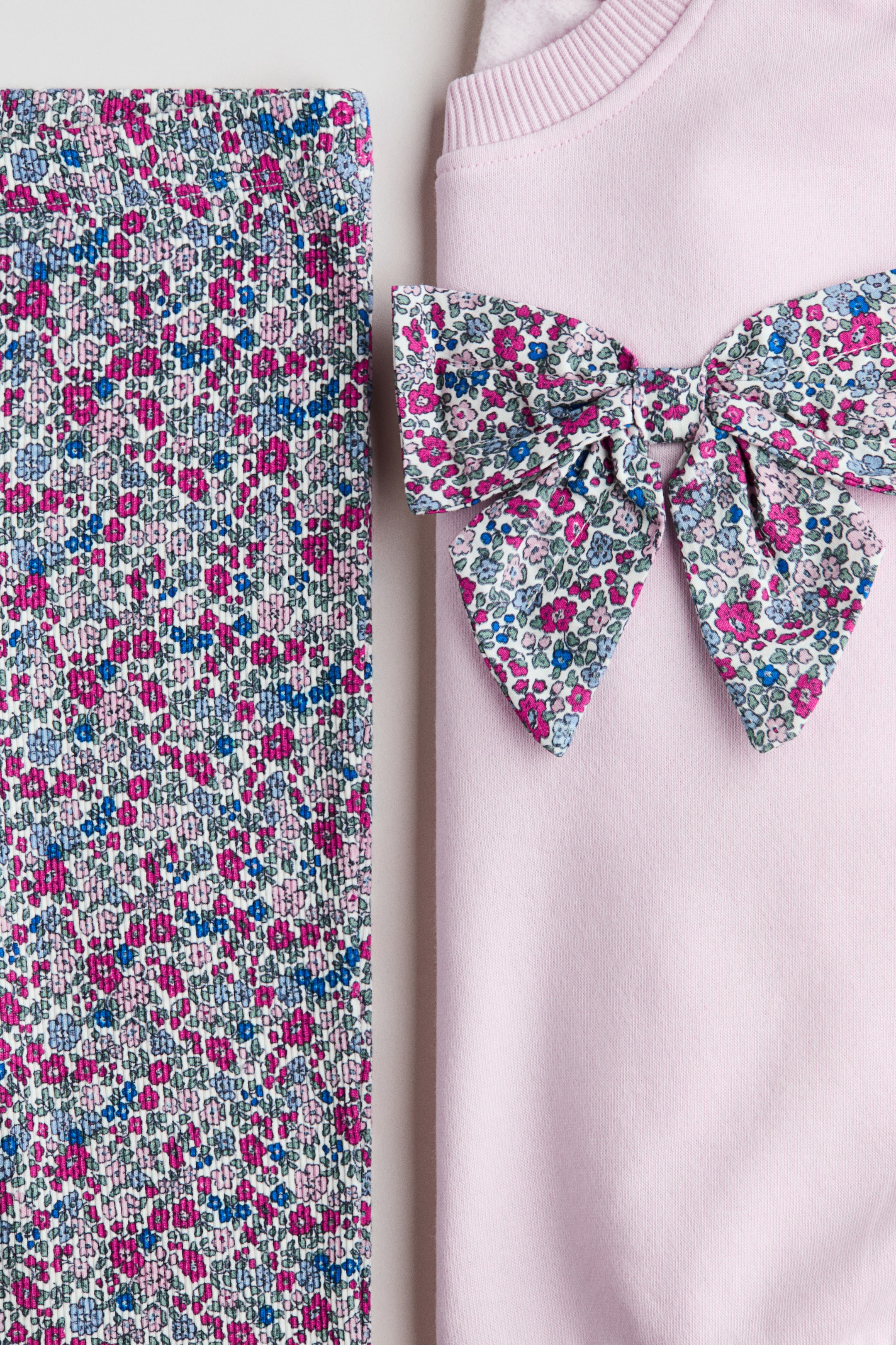 Büyük resmi görüntüle: Açık pembe bir sweatshirt, kendine has bir nervürlü yakası ve ÖNünde beyaz kumaştan yapılmış beyaz üzerine yoğun küçük pembe, mavi ve yeşil çiçek desenli bir çiçek fiyonk ile ön plana çıkıyor. Aynı yoğun küçük çiçek desenini sergileyen uyumlu bir çiçek desenli tayt, yanında katlı halde duruyor.