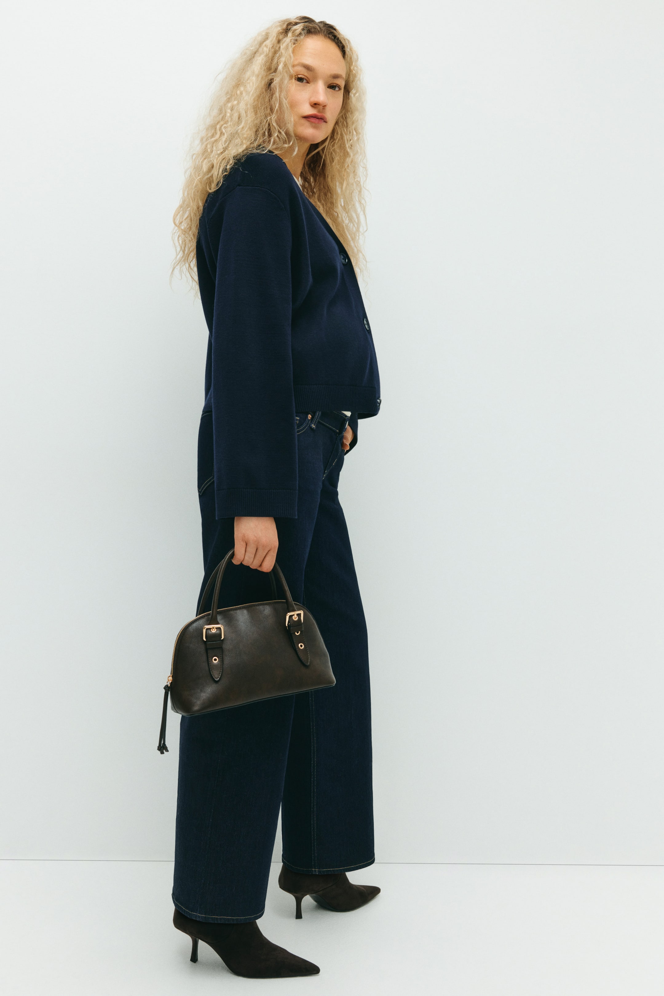View larger image: A dark brown dome-shaped handbag is being held, showing its front and featuring two short top handles, gold-toned decorative buckle details, and a visible zipper closure with a pull tab.