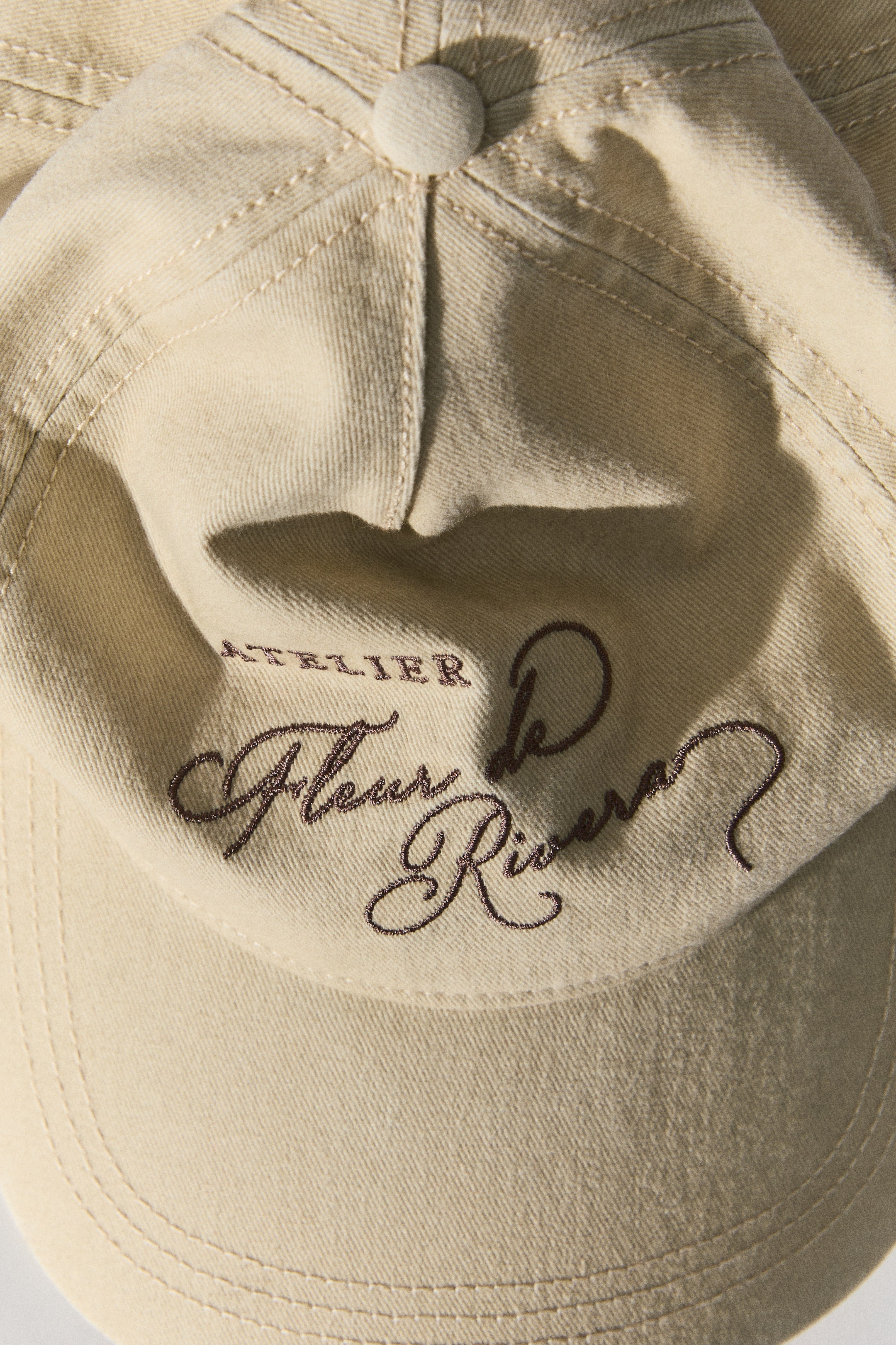 View larger image: A light beige cotton twill cap is shown from the front, displaying dark brown embroidered text that reads 'ATELIER Fleur de Rivera'. The cap features prominent stitching on the crown and brim, with a fabric-covered button visible at the top of the crown.