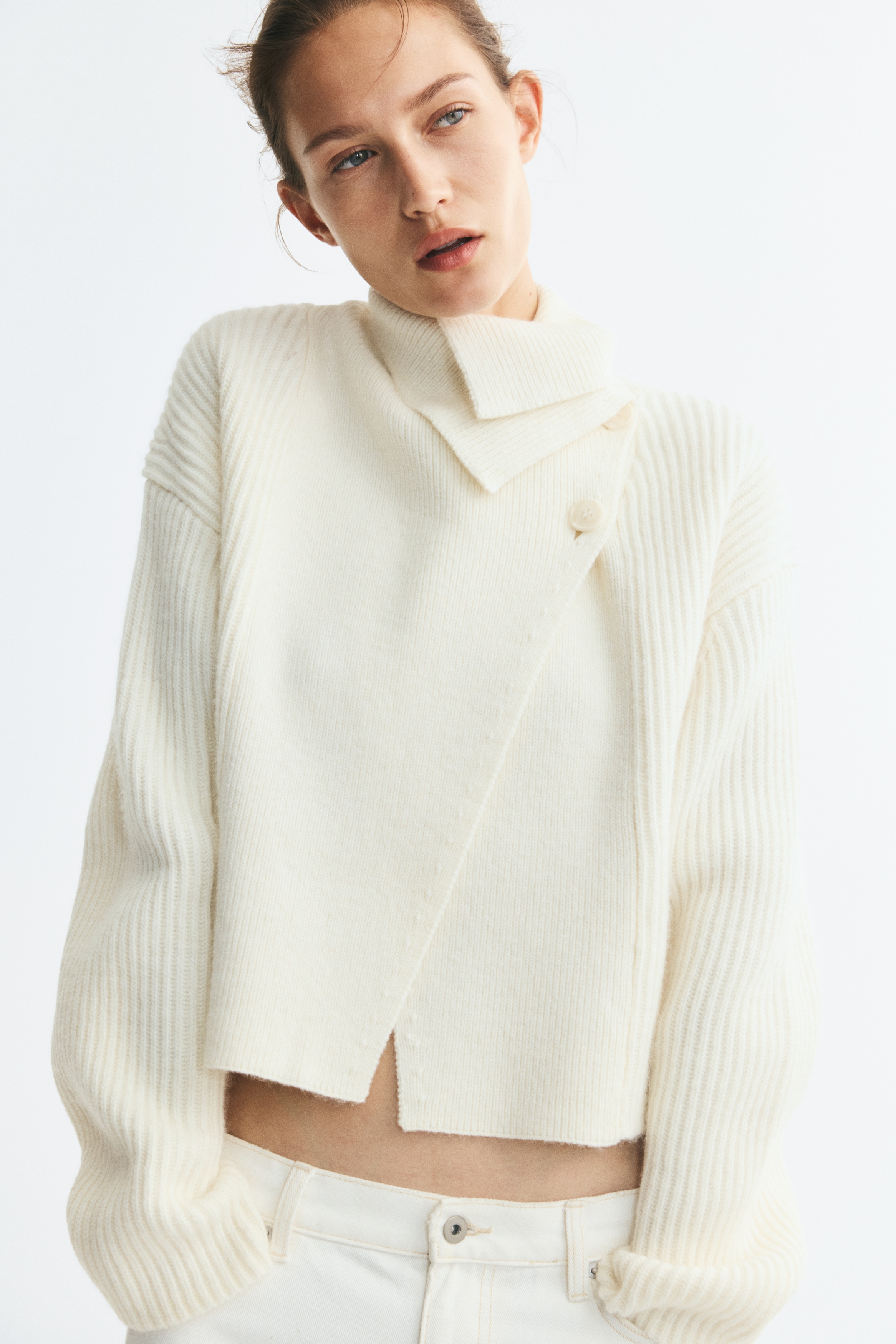 Un pull en maille court blanc cassÃ© est prÃ©sentÃ© de face, avec une fermeture portefeuille asymÃ©trique sur le devant et deux boutons prÃ¨s du col. Le pull prÃ©sente une texture cÃ´telÃ©e sur lâensemble, avec un col montant repliÃ© et de longues manches larges.