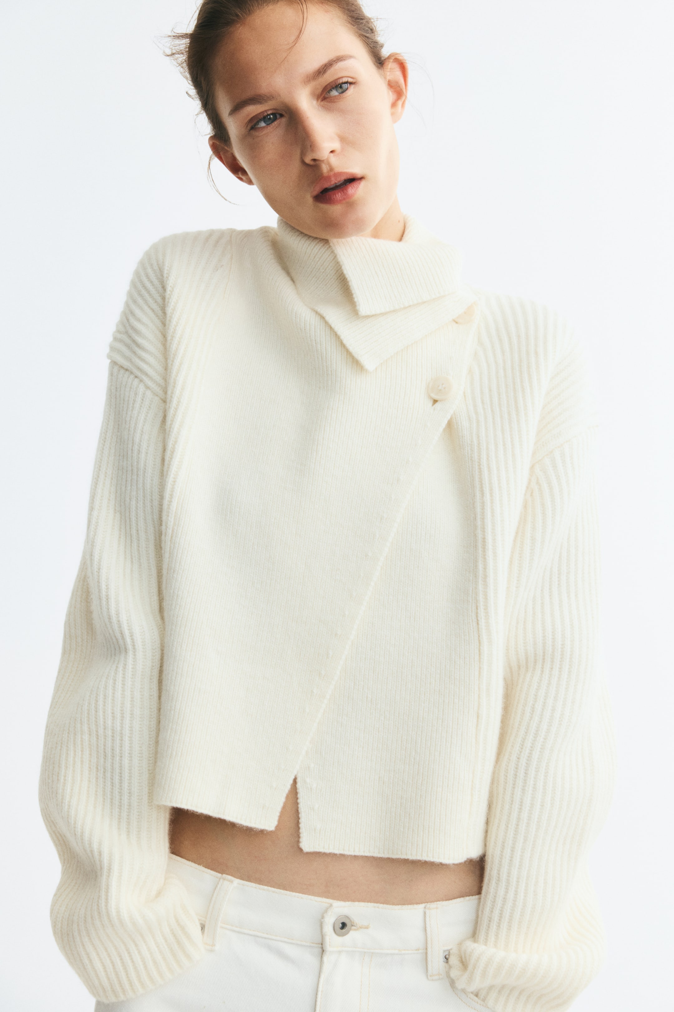 Größeres Bild anzeigen: Ein cremefarbener, kurz geschnittener Cardigan aus Rippstrick wird vom Model getragen und zeichnet sich durch ein Wickeldesign mit Knopfverschluss am Kragen sowie einen hohen, umgeschlagenen Ausschnitt aus. Der Cardigan verfügt über überschnittene Schultern mit langen, voluminösen Ärmeln und ist mit einer weißen Jeans kombiniert.