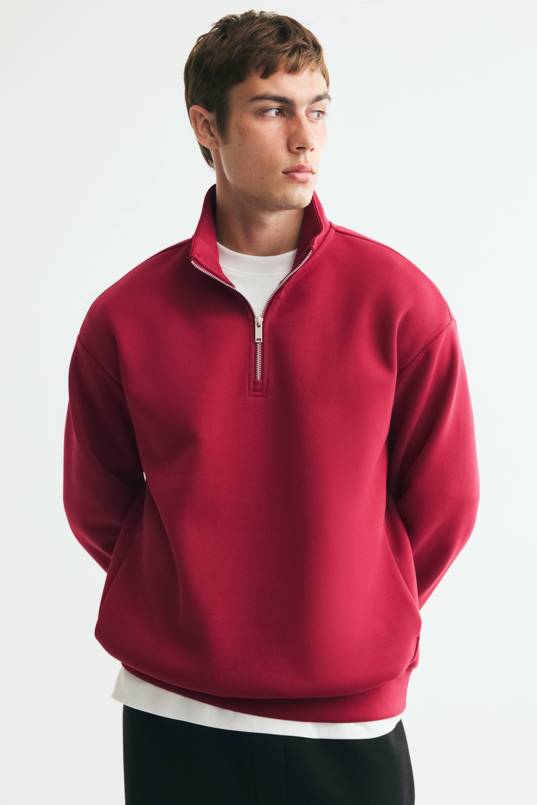 View larger image: The model is wearing a red top made from midweight sweatshirt fabric. It features a stand collar with a partially unzipped silver zipper, softly dropped shoulders, and long sleeves. A white T-shirt is layered underneath the top.