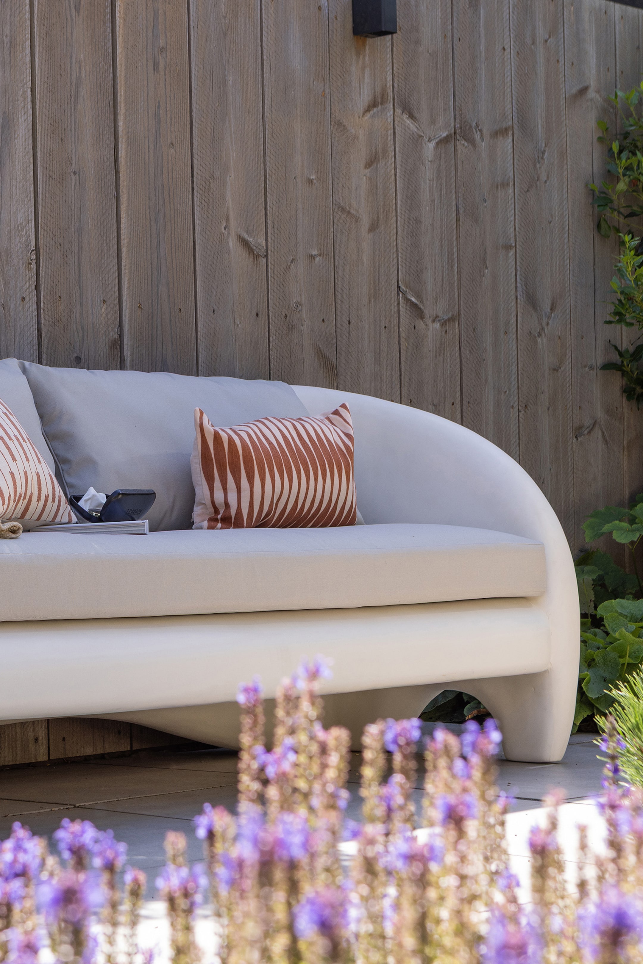 Grotere afbeelding bekijken: Een licht beige gebogen sofa presenteert zijn voor- en rechterzijde, met een solide, afgerond frame en een geïntegreerde, gebogen basis. Het is voorzien van een licht beige zitkussen, twee lichtgrijze rugkussens en twee sierkussens met een golvend roodbruin streeppatroon.