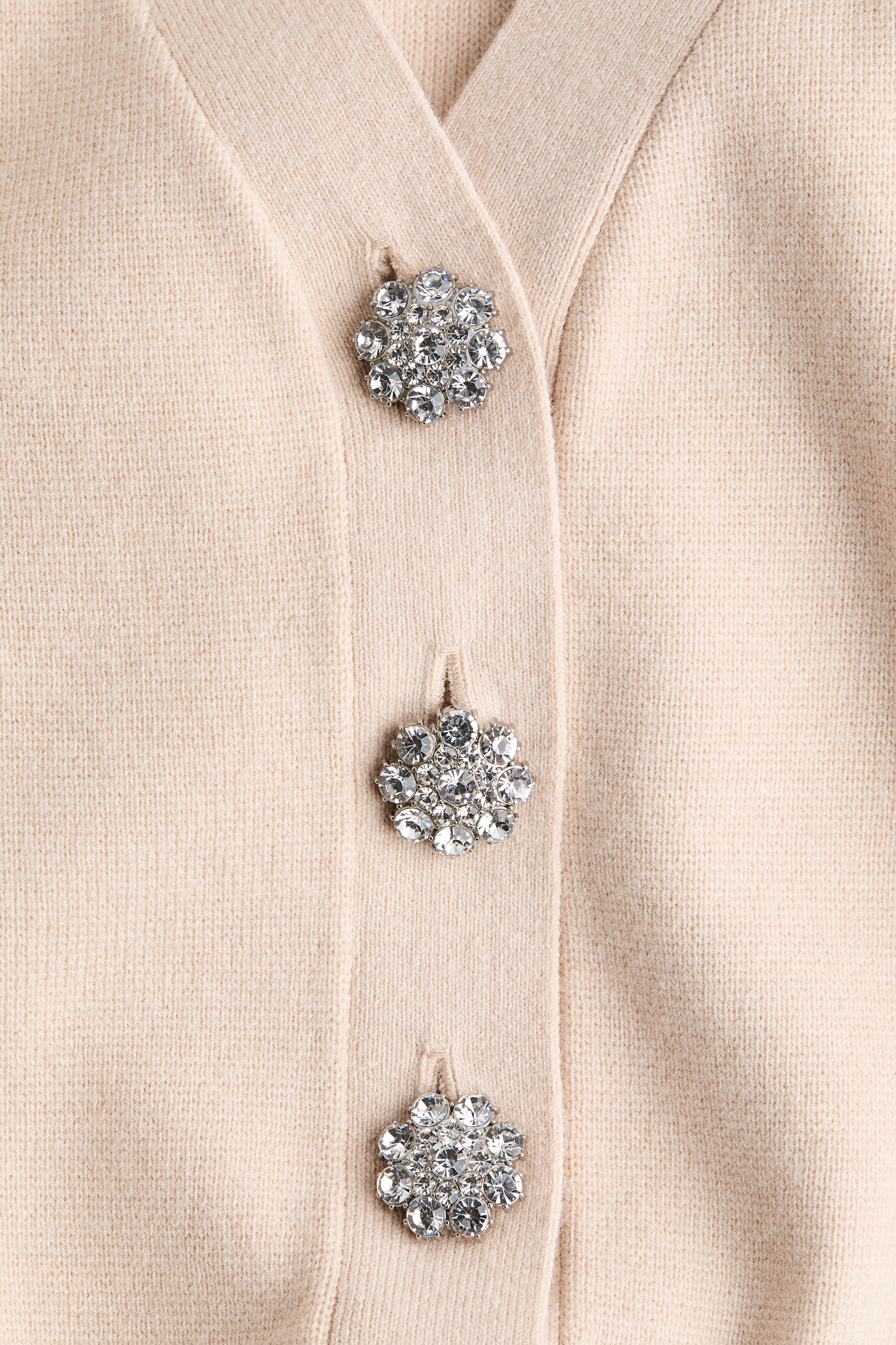 Ampliar la imagen: Un cárdigan de punto suave en color beige claro presenta un escote en V y una hilera de botones frontales. Está adornado con tres botones visibles en forma de flor, cada uno mostrando múltiples cristales transparentes.