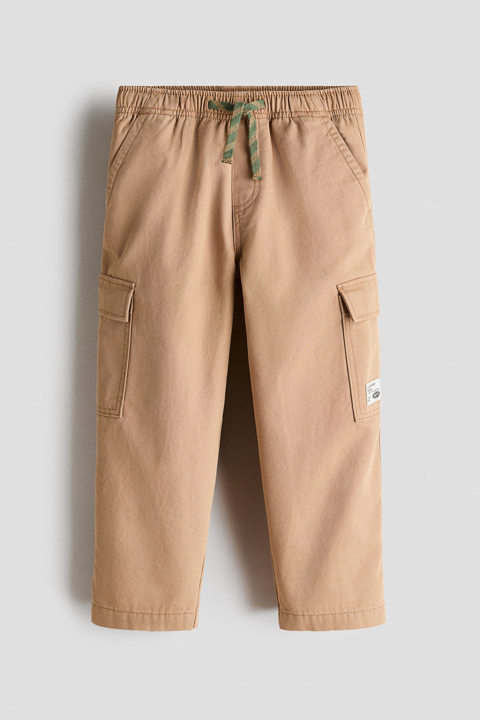 Tan cargo pants are laid flat, featuring an elasticated waistband with a green and brown patterned drawstring. They are showcasing front slant pockets and side cargo pockets with flap closures, with a small white label visible on the right cargo pocket. - 1