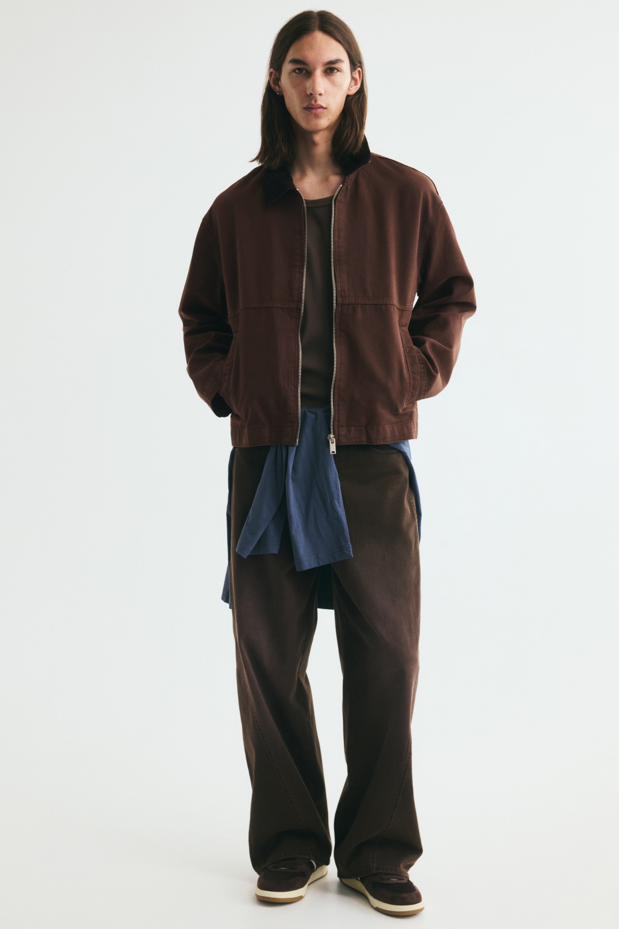 View larger image: The dark brown short workwear jacket in cotton canvas, featuring a partially unzipped front and a contrasting dark corduroy collar, is being worn by the model, who is placing their hands into its welt front pockets. A brown T-shirt is visible underneath, with a blue shirt tied around the waist.