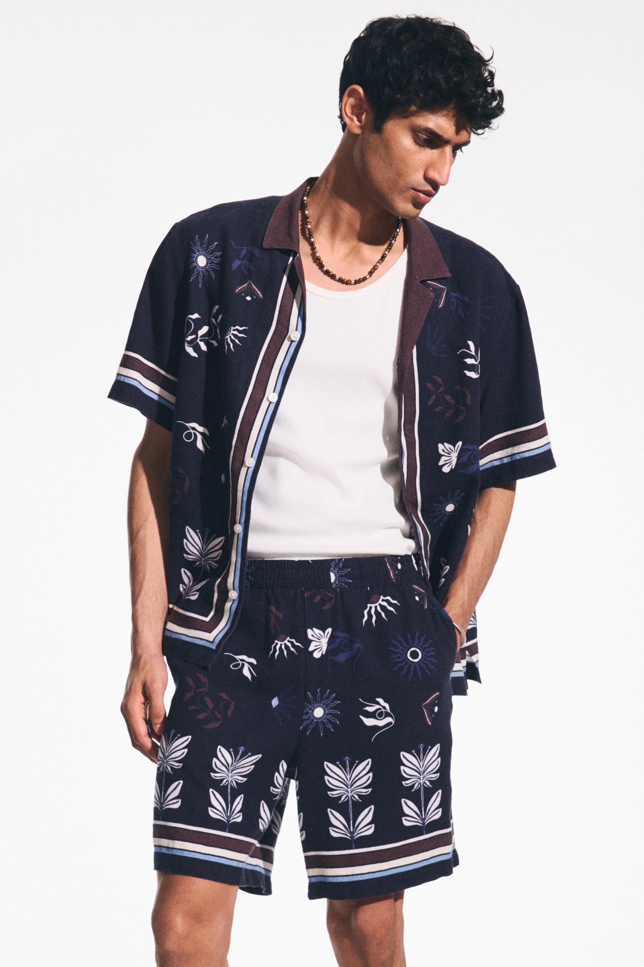 View larger image: A navy blue short-sleeved shirt and matching shorts set features an all-over abstract pattern in white, light blue, and burgundy. The shirt showcases a burgundy camp collar with vertical stripe detailing on the placket and sleeve cuffs, worn open over a white t-shirt. The shorts come with an elasticated waistband and coordinating horizontal stripe detailing near the hem.