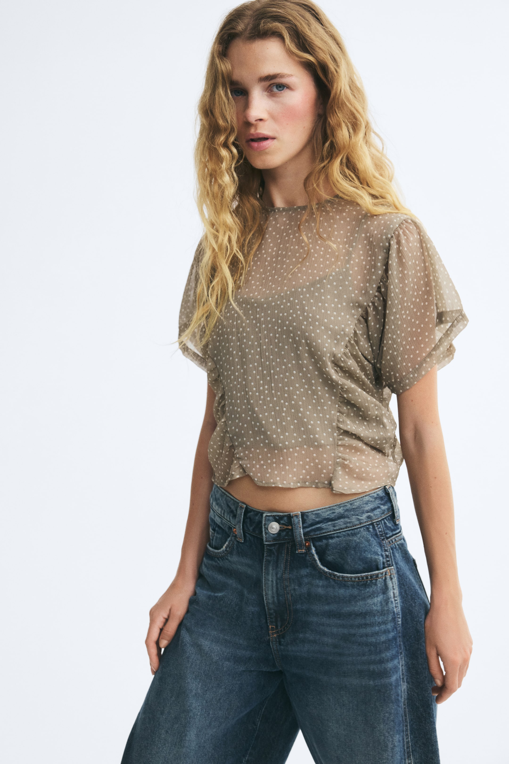 View larger image: The model is wearing a sheer chiffon blouse in a taupe polka-dot print, featuring a round neckline, short flutter sleeves, and gathered side panels. The blouse is layered over a dark underlayer and paired with blue jeans.