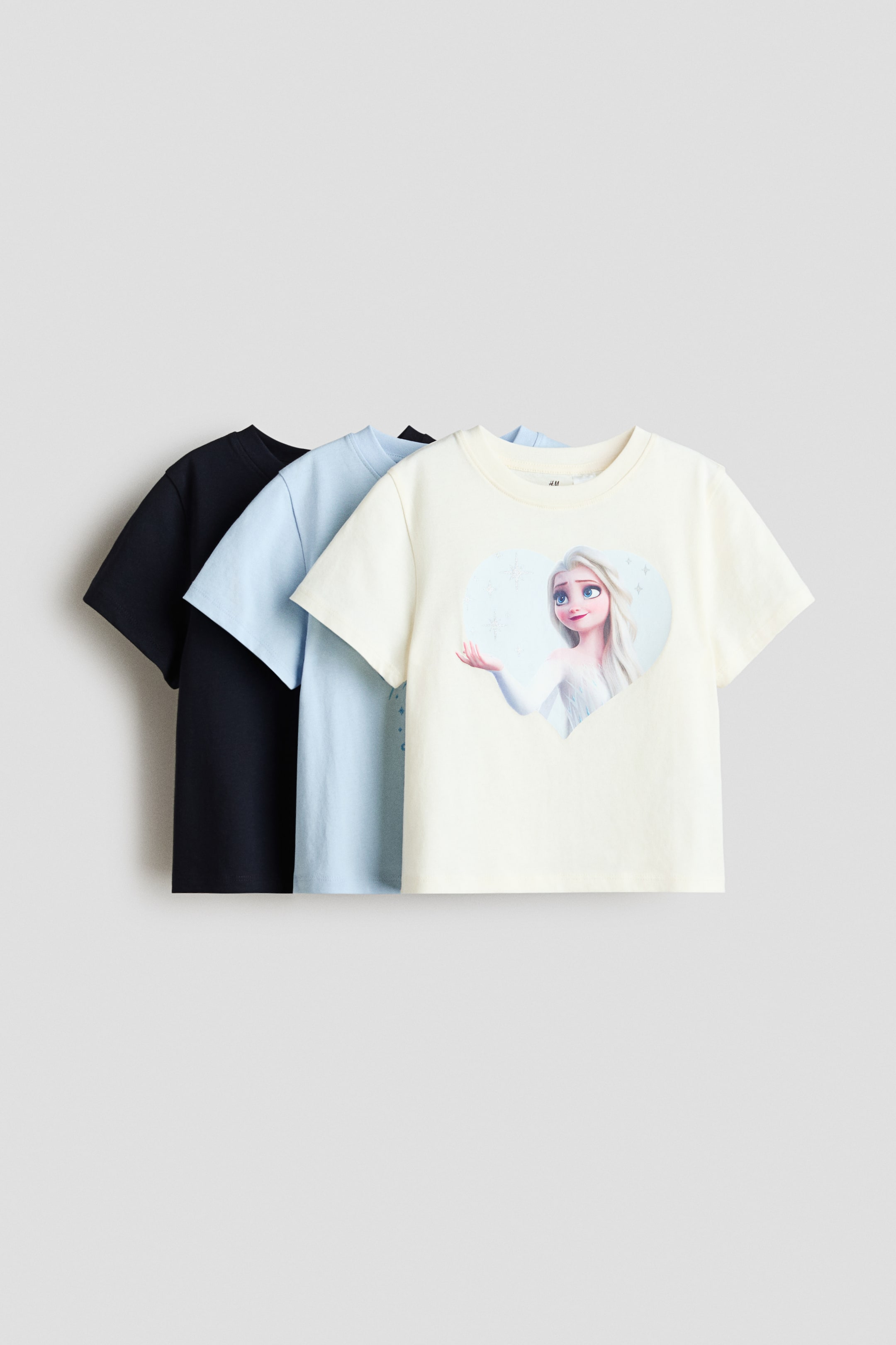 View larger image: Three short-sleeved tops crafted from soft cotton jersey are displayed from the FRONT, giving a layered look in cream, light blue, and dark blue. The cream top features a print motif of Elsa from Frozen inside a heart shape on its front.