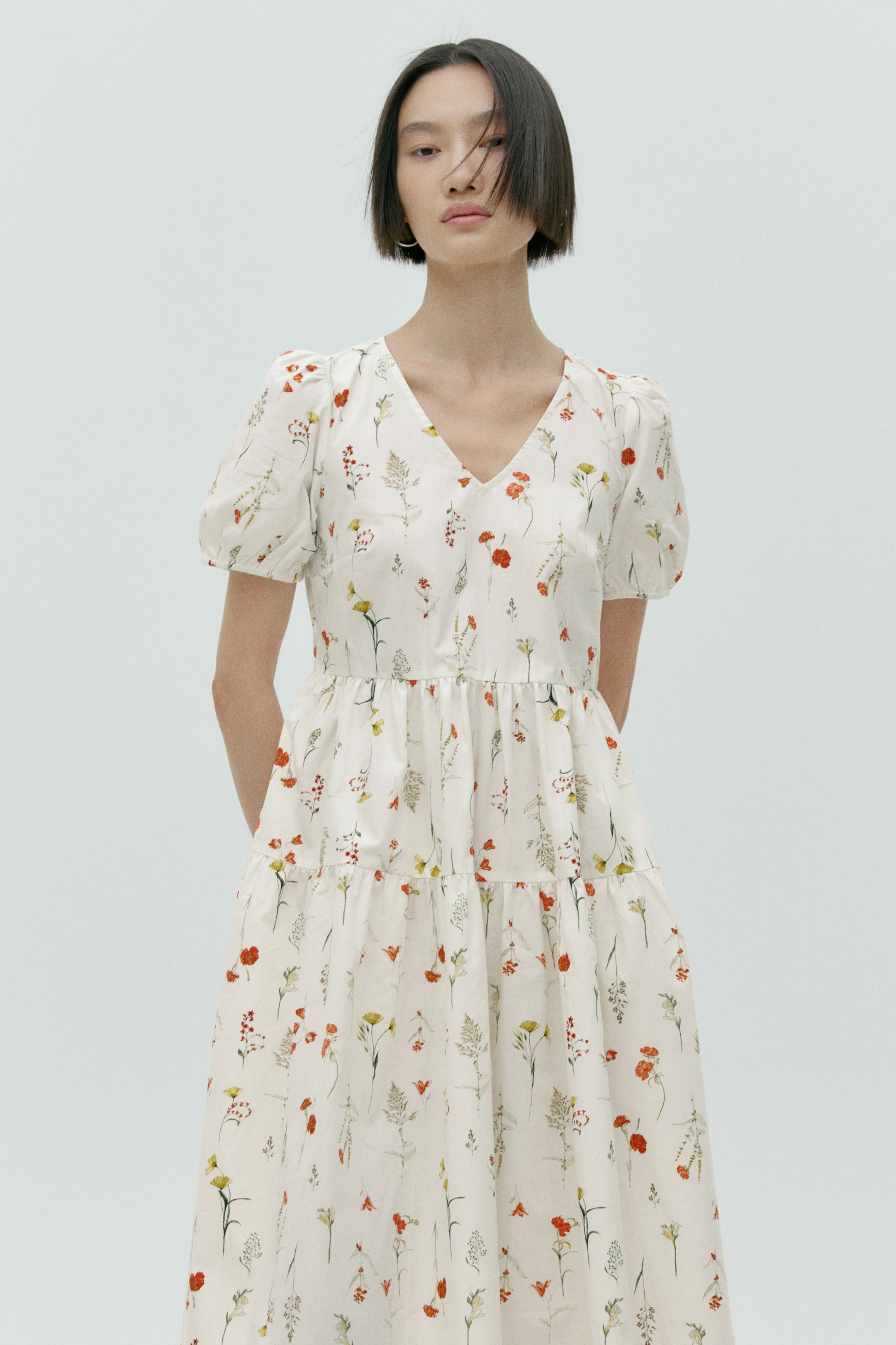 View larger image: A white dress features a V-neckline and short puffed sleeves, showcasing a scattered print of red, yellow and green wildflowers, with a fitted bodice that transitions into a tiered, flowing skirt.