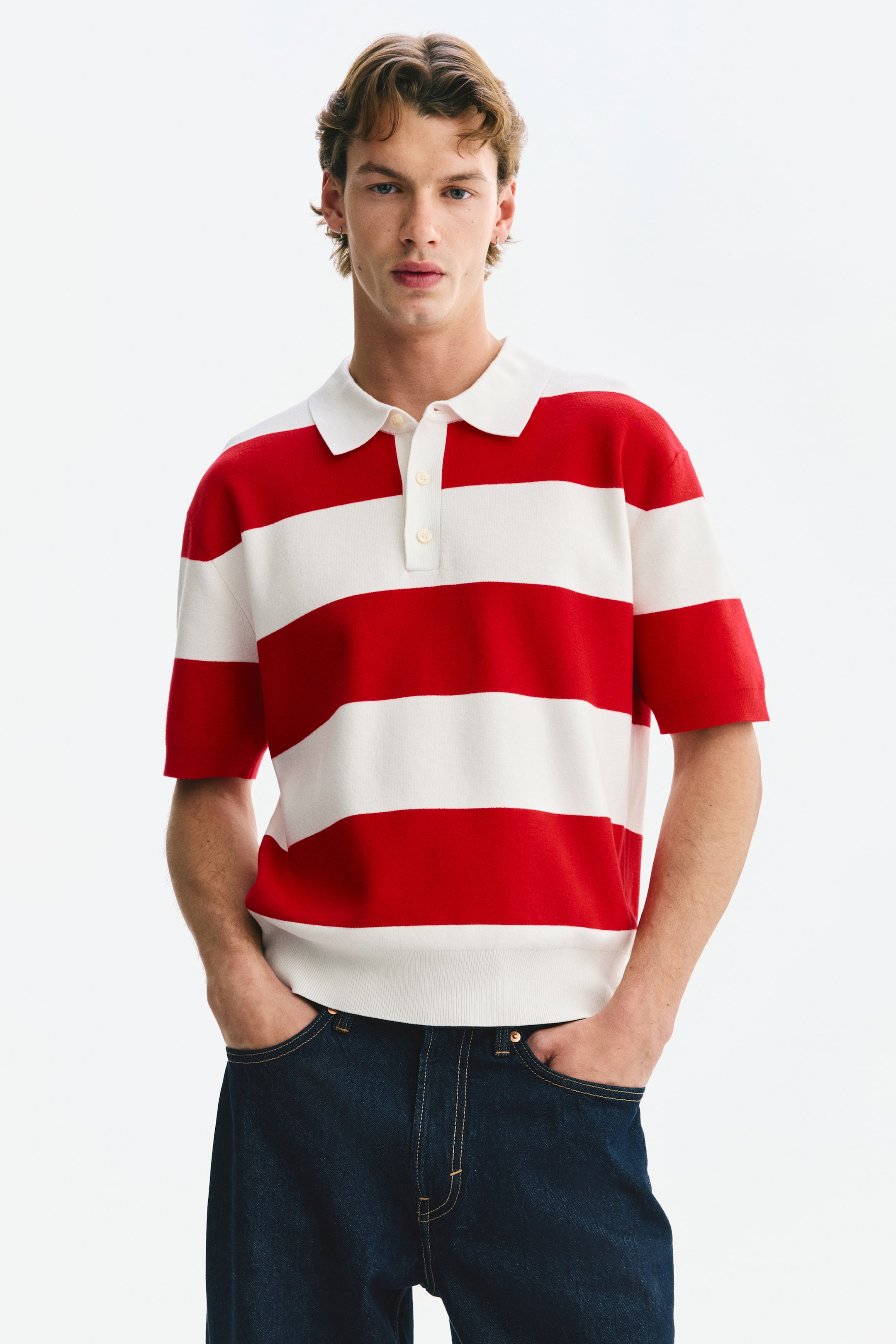 View larger image: The model is wearing a red and white horizontally striped knit polo shirt. It features a white collar, a three-button placket, short sleeves with ribbed cuffs, and a ribbed hem. The regular fit polo shirt is paired with dark denim jeans.