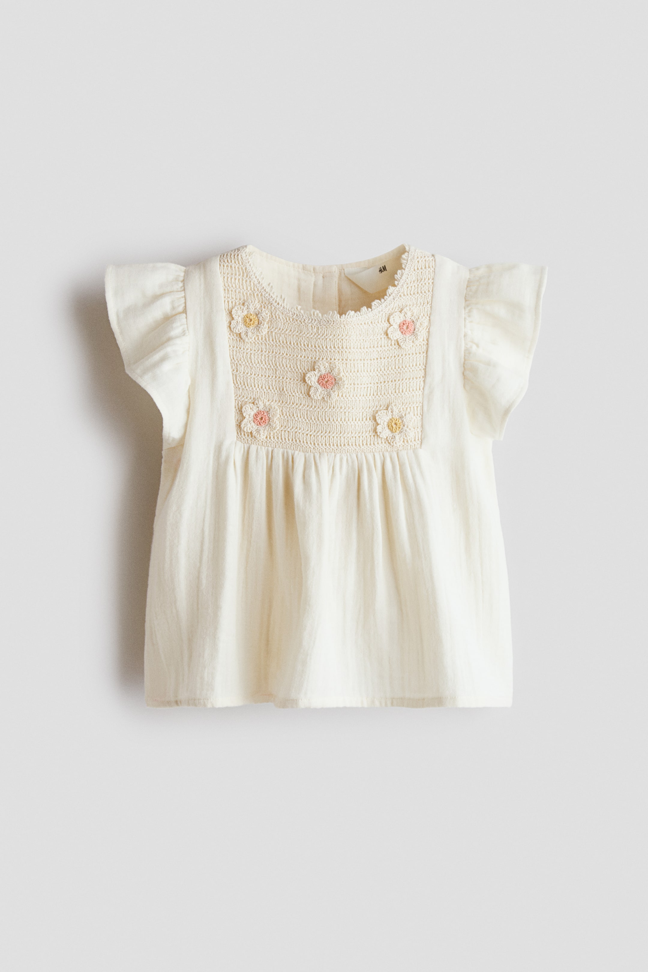 View larger image: A cream-coloured baby top showcases a square, crocheted yoke on its front, adorned with small embroidered flowers in shades of pink and yellow. Ruffled cap sleeves frame the shoulders, while the main body features a soft, crinkled texture.