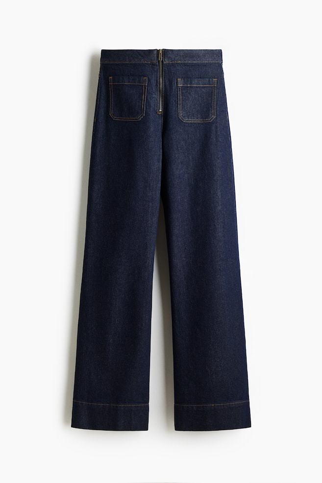 Donkerblauwe denim jeans worden van de achterkant getoond, met een extra-hoge taille en een zichtbare messingkleurige ritssluiting aan de achterkant. Ze zijn ontworpen met twee rechthoekige opgestikte zakken aan de achterkant en wijde pijpen die tot aan de zoom reiken, allemaal geaccentueerd door lichtbruine contraststiksels. - 1