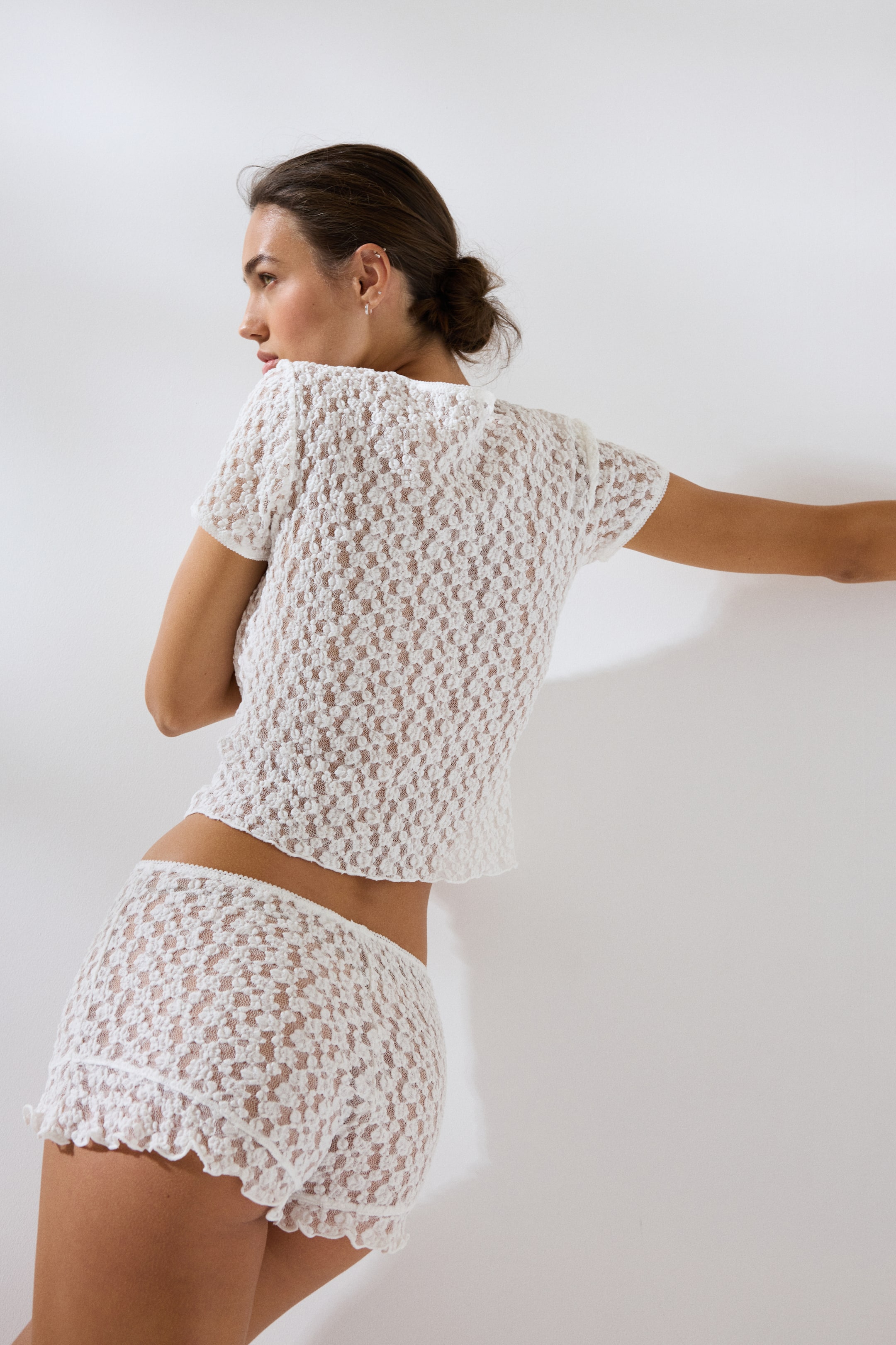 View larger image: A white crochet knit two-piece set, viewed from a back-side angle, features a short-sleeved top and matching shorts. Both pieces showcase an open-weave pattern and scalloped trim along the top's hem and sleeve cuffs, as well as the shorts' leg openings.