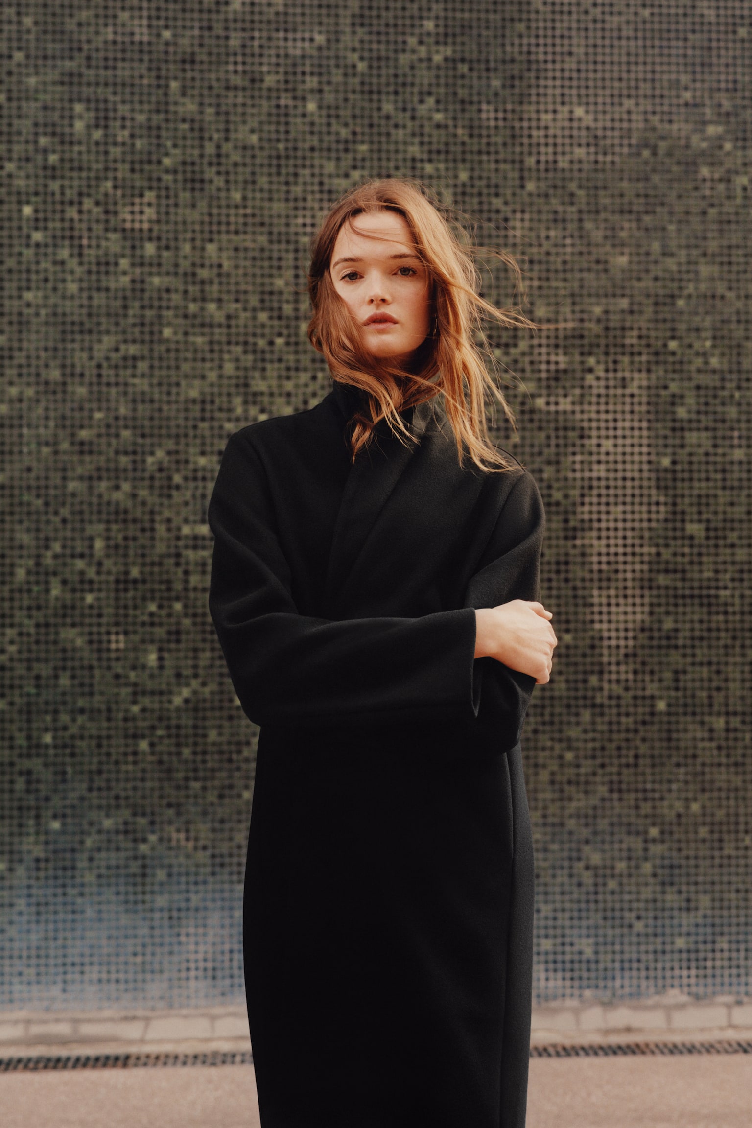 A black, knee-length coat in a soft, felted fabric is worn by the model, who is standing with arms crossed. The coat features long, relaxed sleeves and is styled with a high, wrapped front closure. - 1